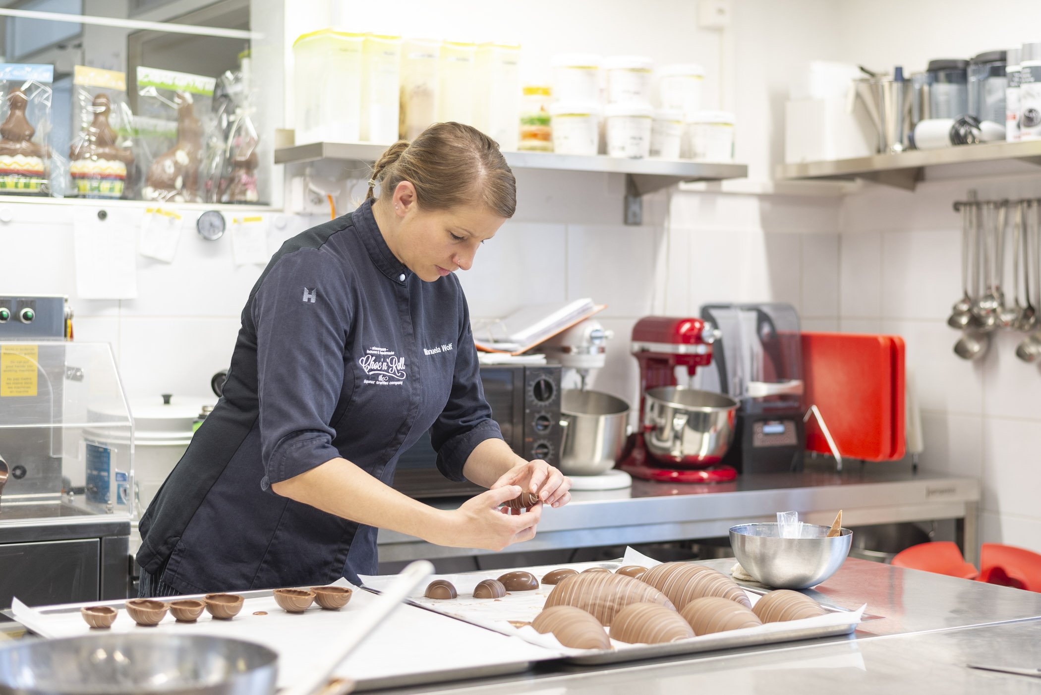 Frau in einer Bäckerei oder Konditorei, die Schokoladenformen auf einem Tisch herstellt. Im Hintergrund sind Küchengeräte und -utensilien zu sehen, darunter ein Mixer und diverse Behälter.