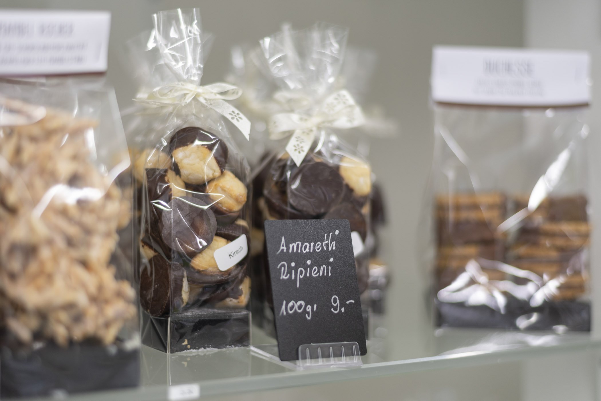 Verpackte Amaretti Ripieni und Kekse hinter einer Glasscheibe, mit einem Preisschild "Amaretti Ripieni 100g 9.-" in einem Geschäft.