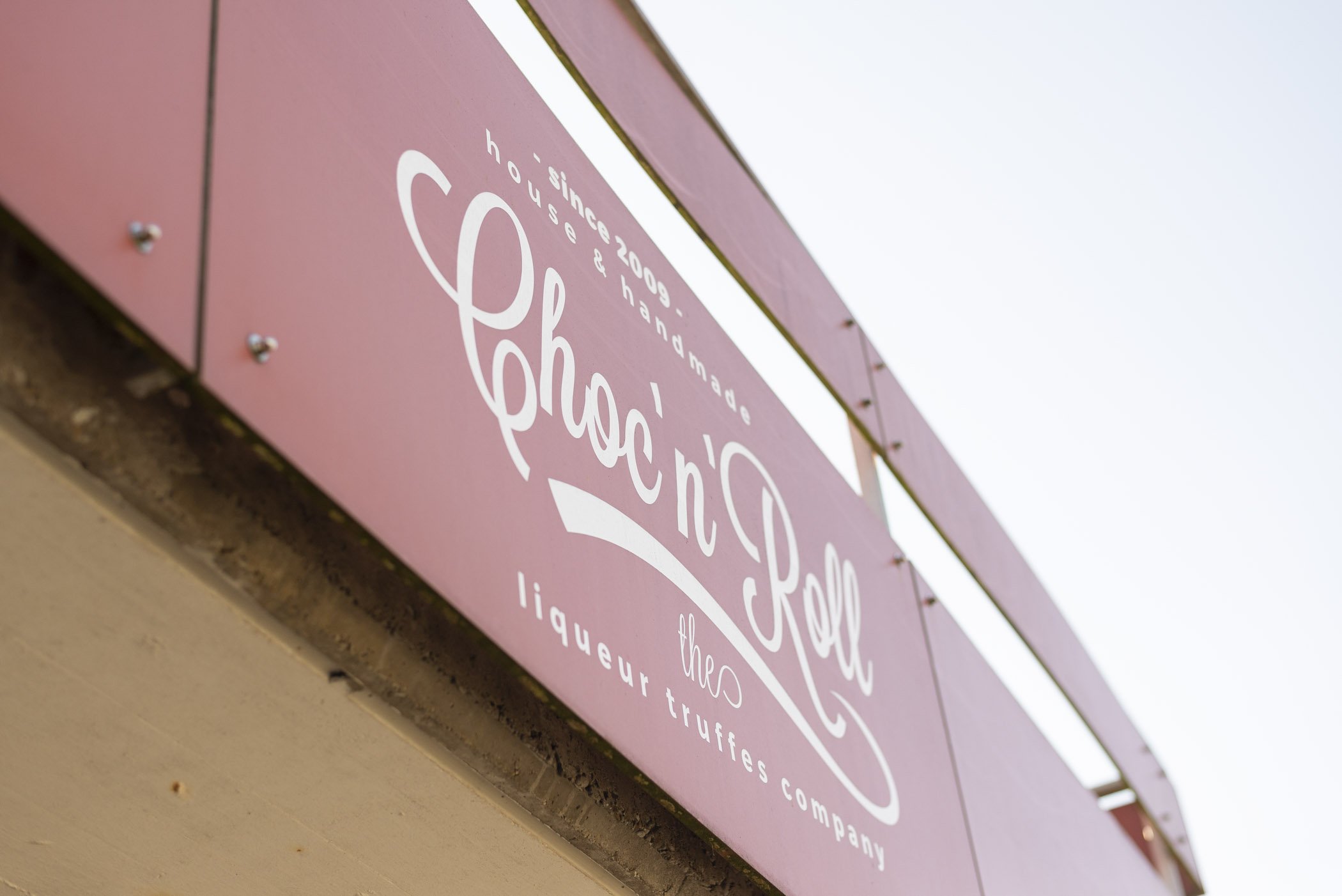 Nahaufnahme eines rosa Schildes mit der Aufschrift "Choc'n'Roll" und dem Untertitel "the liqueur truffles company", darunter steht "since 2009, house & handmade".