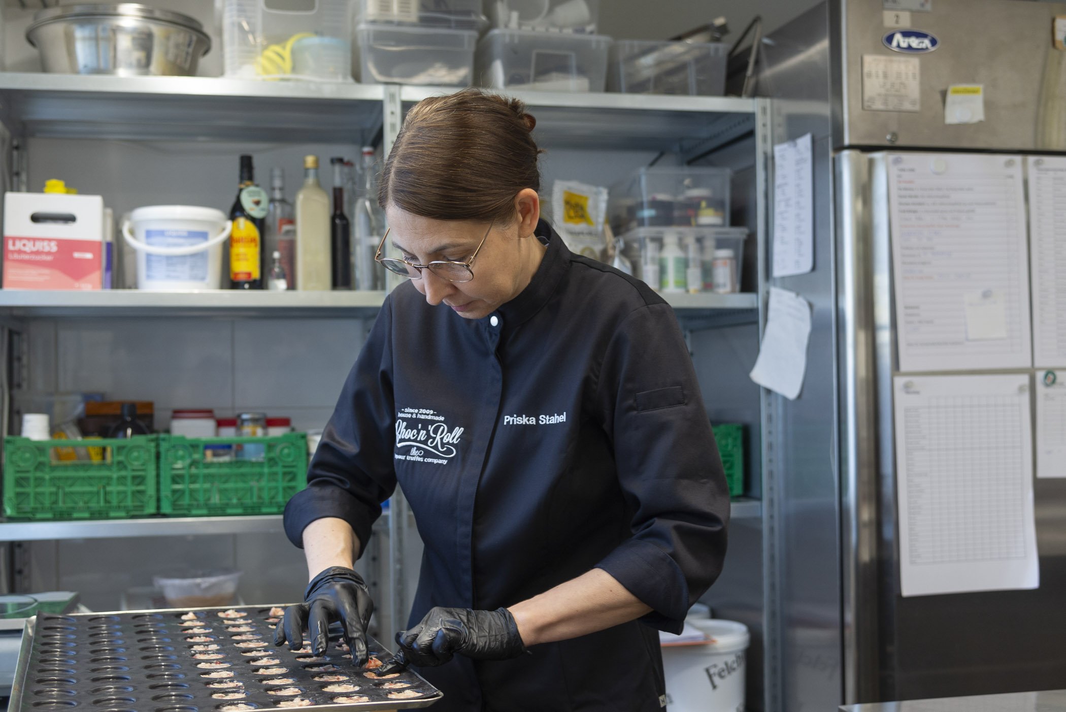 Eine Frau in schwarzer Küchenuniform arbeitet in einer modernen Küchenumgebung und formt Schokolade in einer Form. Im Hintergrund sind Regale mit Zutaten und Utensilien zu sehen.