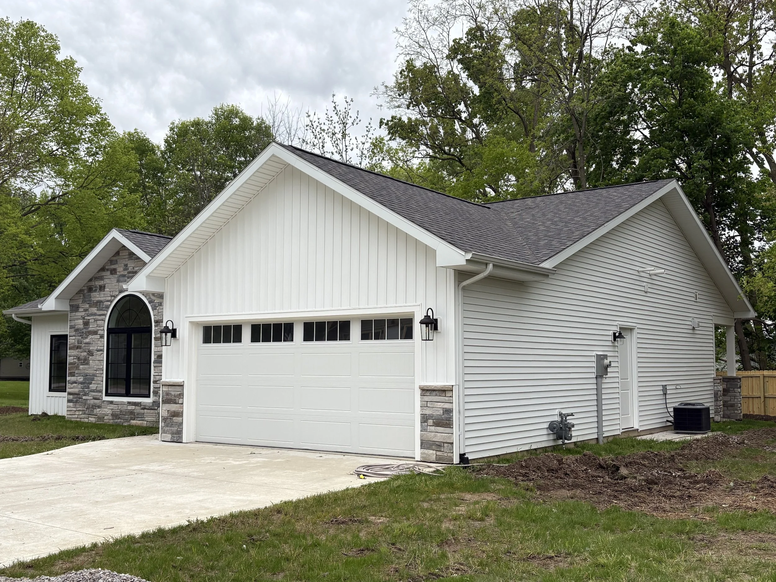 A new white house with gray shingled roof, stone accents, and a garage door, surrounded by green trees and a cloudy sky.