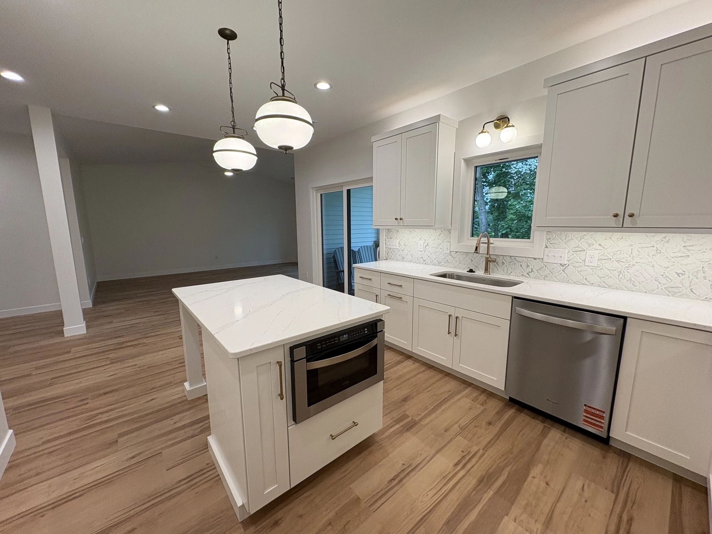 Modern kitchen with white cabinets, a marble island, stainless steel appliances, pendant lighting, and a window overlooking a backyard with trees.