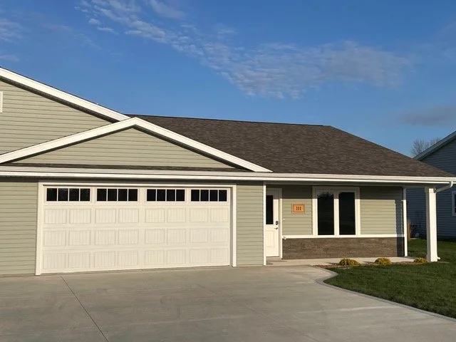 Front view of a single-story house with an attached garage, green siding, white trim, and a small front yard.