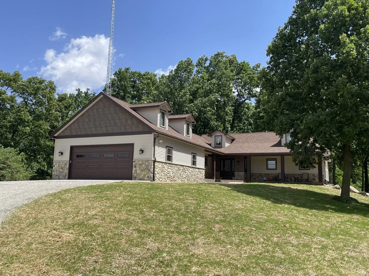 A two-story house with a brown roof, stone foundation, beige siding, and a two-car garage, surrounded by green trees on a sunny day.