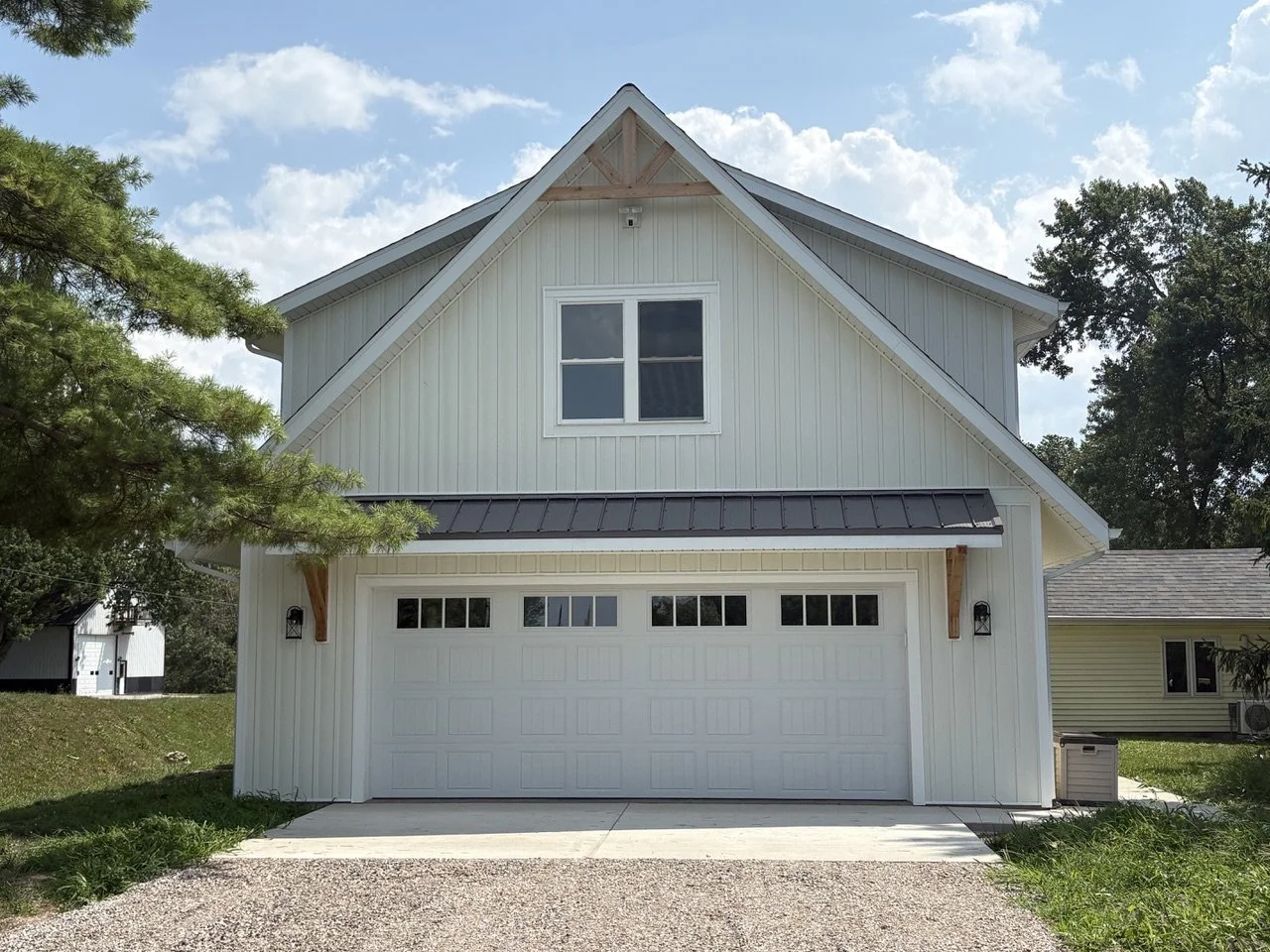 A modern two-story white house with a garage door, a window above the garage, and a metal roof. There are trees and a partly cloudy sky in the background.