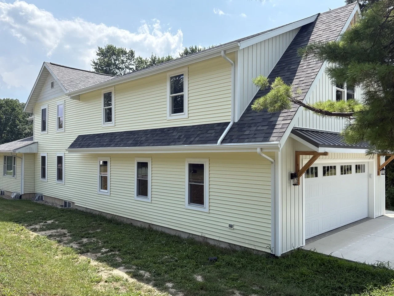 A two-story house with light yellow vinyl siding, multiple white-framed windows, a black shingled roof, and a white garage door. There is a concrete driveway and some grass in the front yard, with a tree extending into the frame from the right.