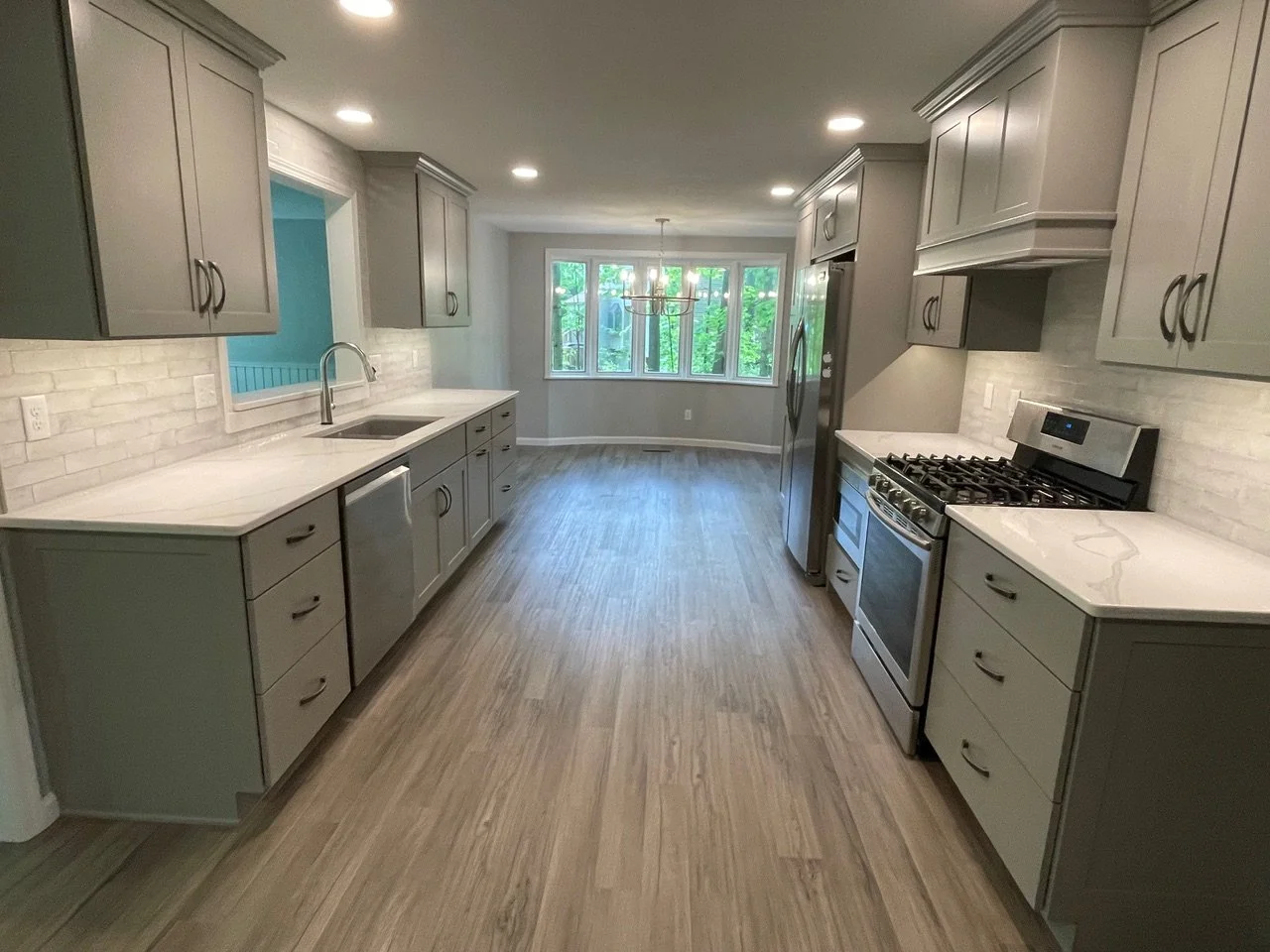 Modern kitchen with gray cabinets, white countertops, stainless steel appliances, and hardwood floors, with a bay window at the far end.