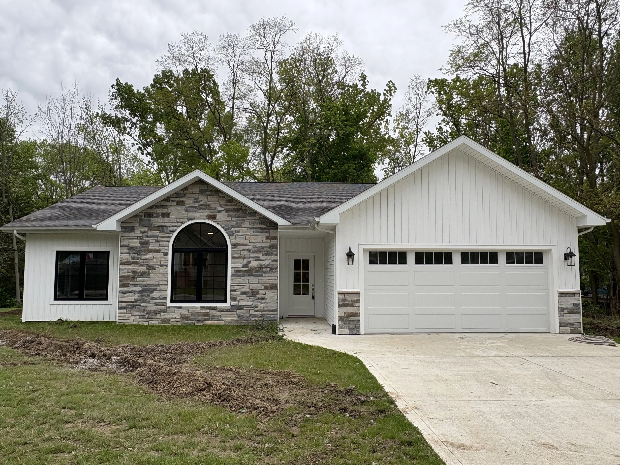 Front view of a new house with white siding, a stone accent wall near the entrance, a garage, and a driveway. Trees are visible in the background and the sky is overcast.