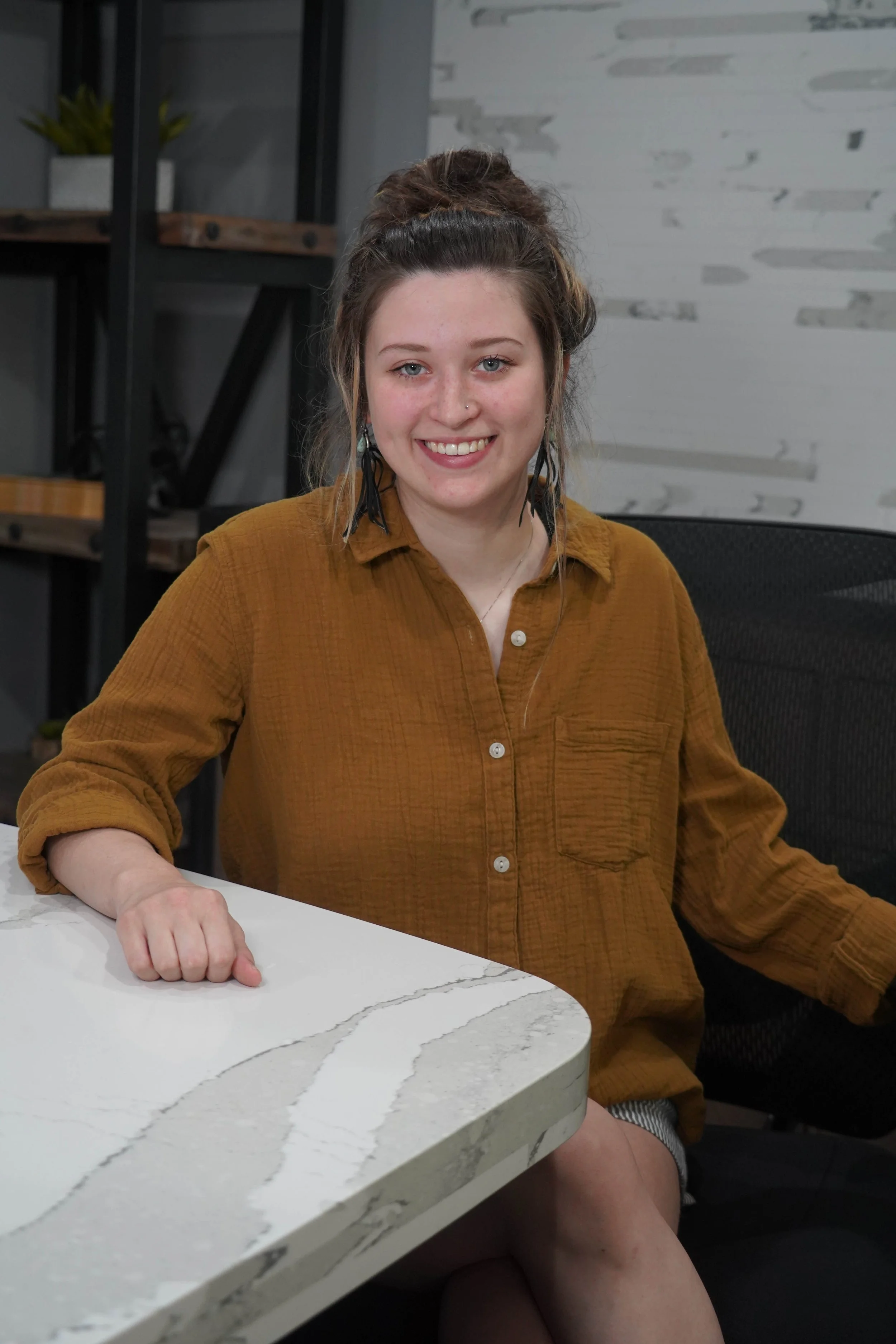A young woman with light skin, blue eyes, and ear piercings, smiling, sitting at a white marble table in a modern office or kitchen space, wearing a mustard-colored button-up shirt, with her hair in a messy bun.
