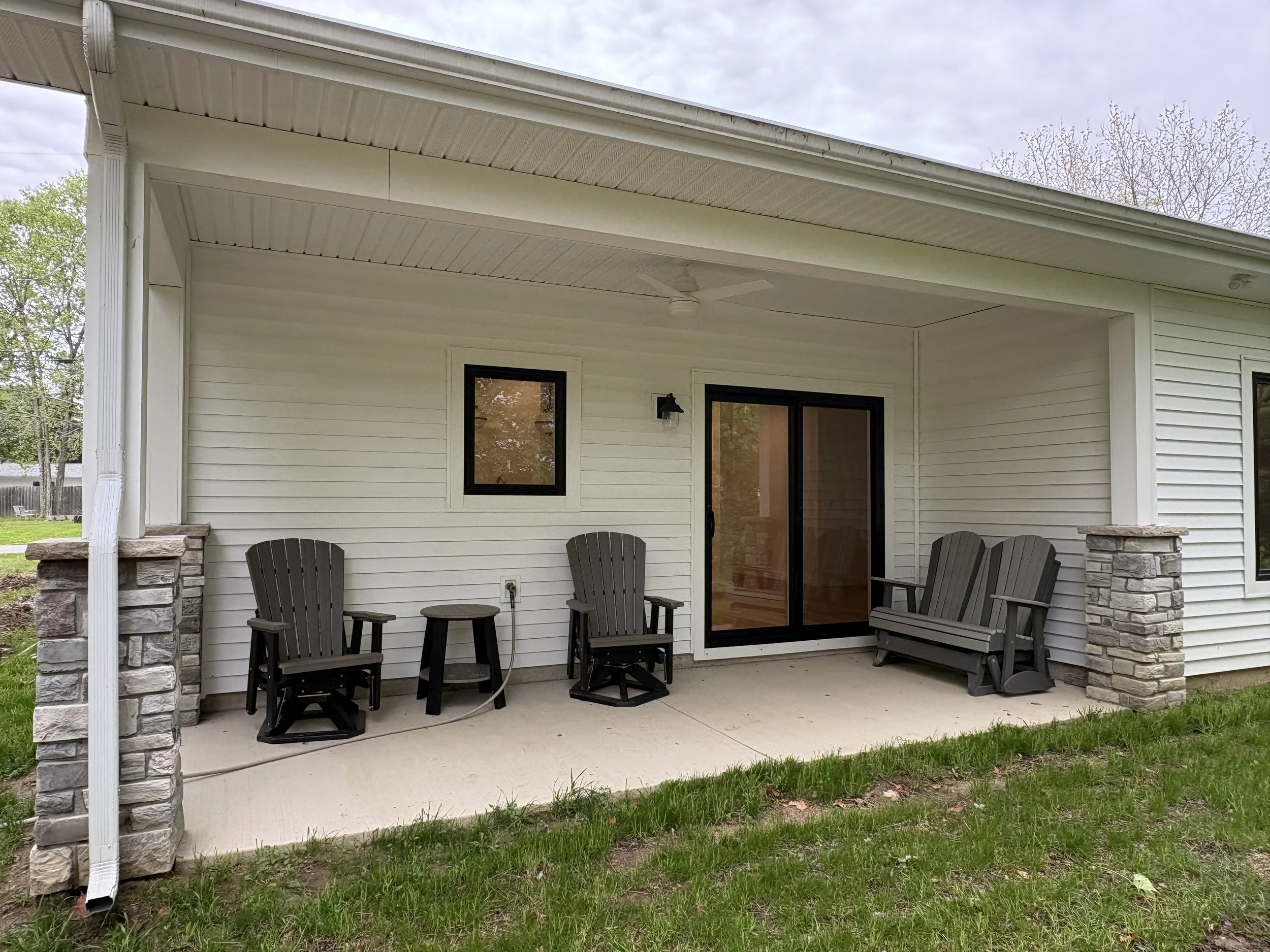 A covered outdoor patio area attached to a house, with four chairs and a small table, a ceiling fan, a wall-mounted lantern, and sliding glass doors.