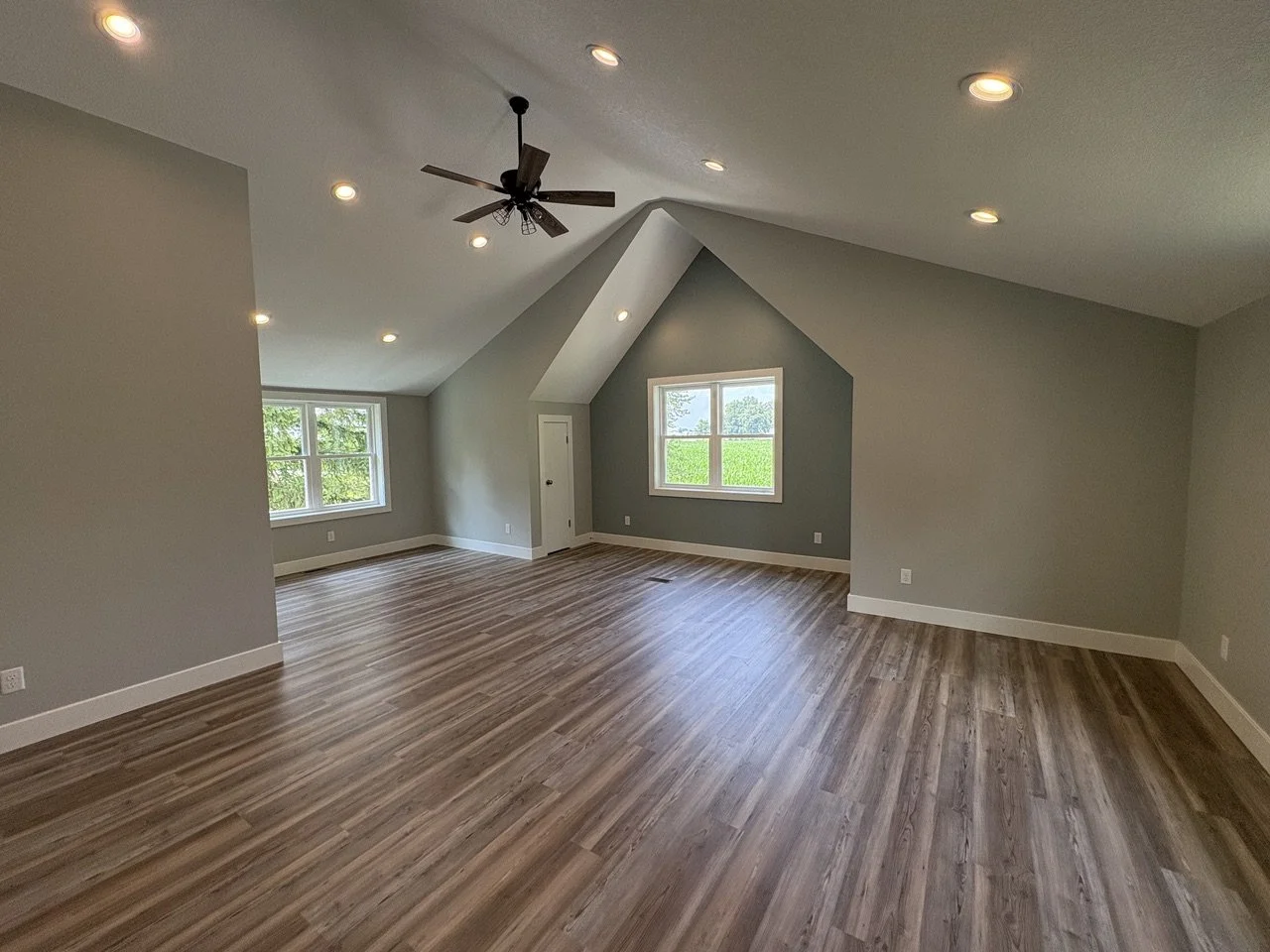 Empty room with sloped ceiling, wood flooring, gray walls, four ceiling lights, and two windows showing green trees outside.