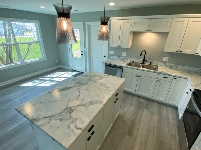 Modern kitchen with white cabinets, marble countertops, a kitchen island, and stainless steel appliances. Two pendant lights hang above the island, with large windows providing natural light.
