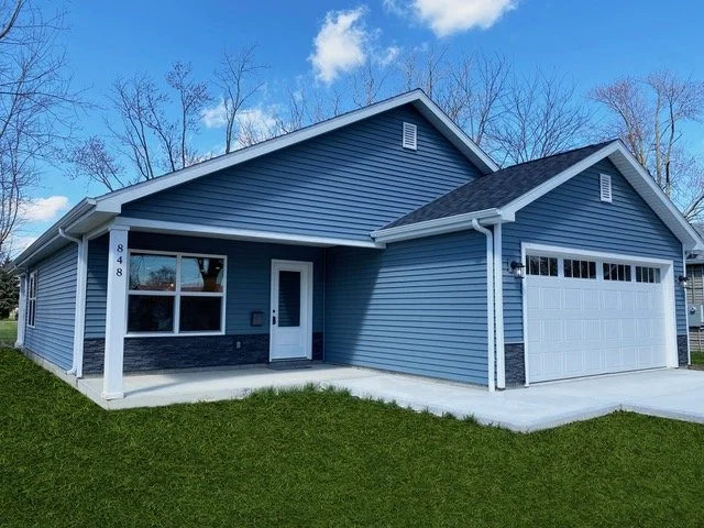 A blue house with a white garage door, white trim, and black accents. The house has a front lawn and a concrete walkway leading to the front door. The sky is partly cloudy.