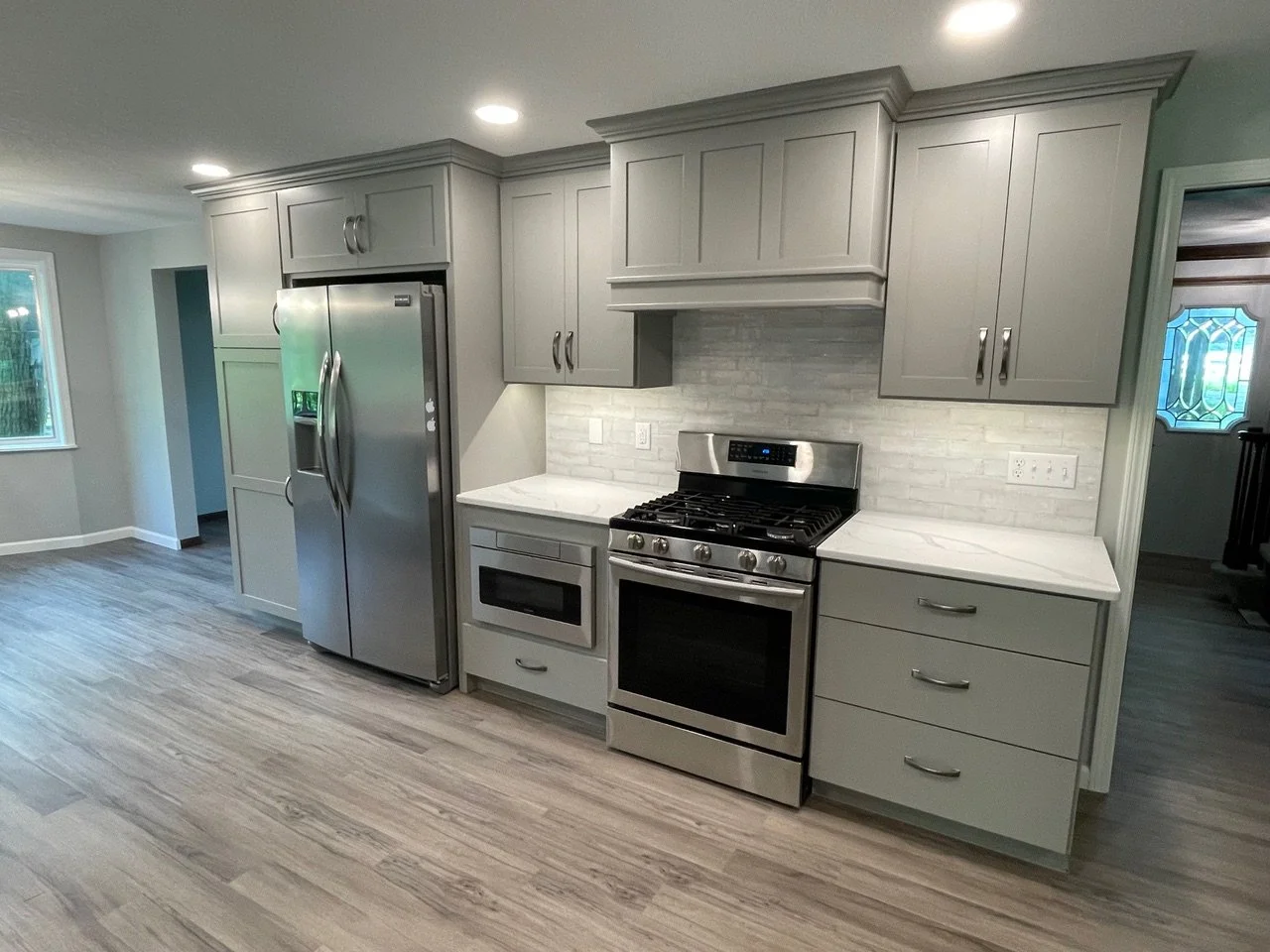 Modern kitchen with gray cabinets, stainless steel refrigerator and oven, white marble countertops, gray brick backsplash, and wood-look flooring.