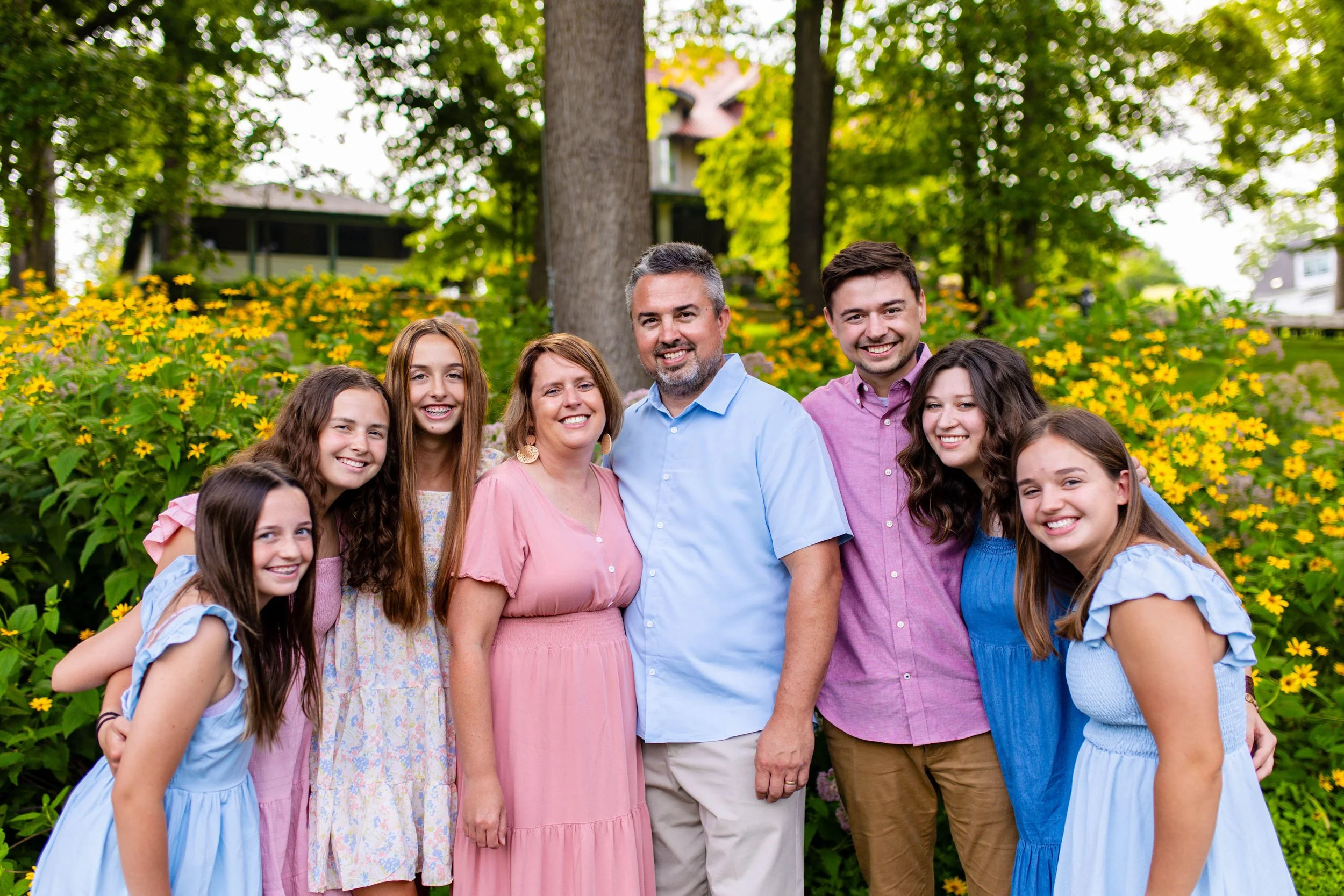 A family group, consisting of three adult men and three young girls, standing outdoors in front of a garden with yellow flowers and green trees, smiling at the camera.