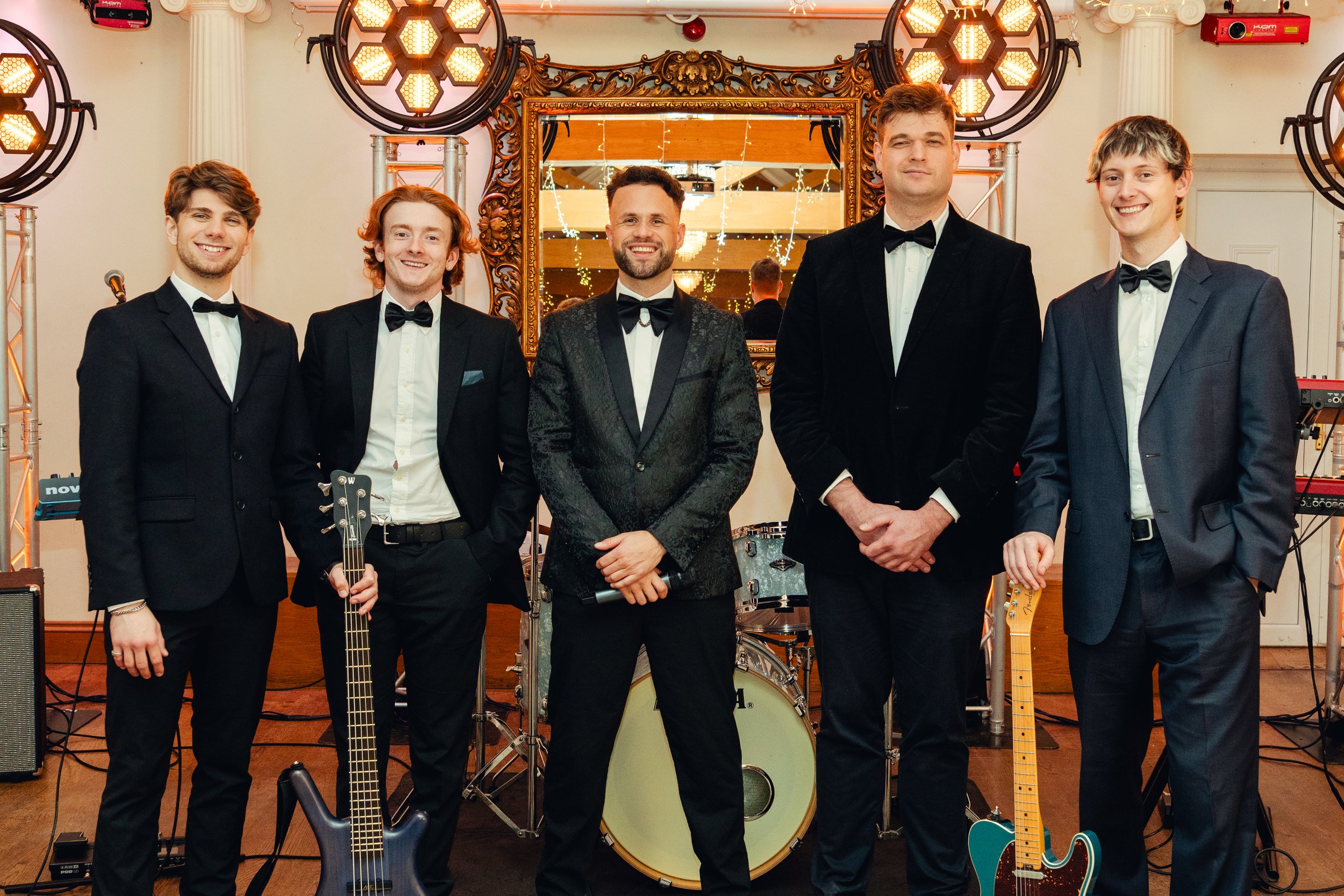 Five musicians in formal attire standing with instruments in a bright room with large windows and skylight.