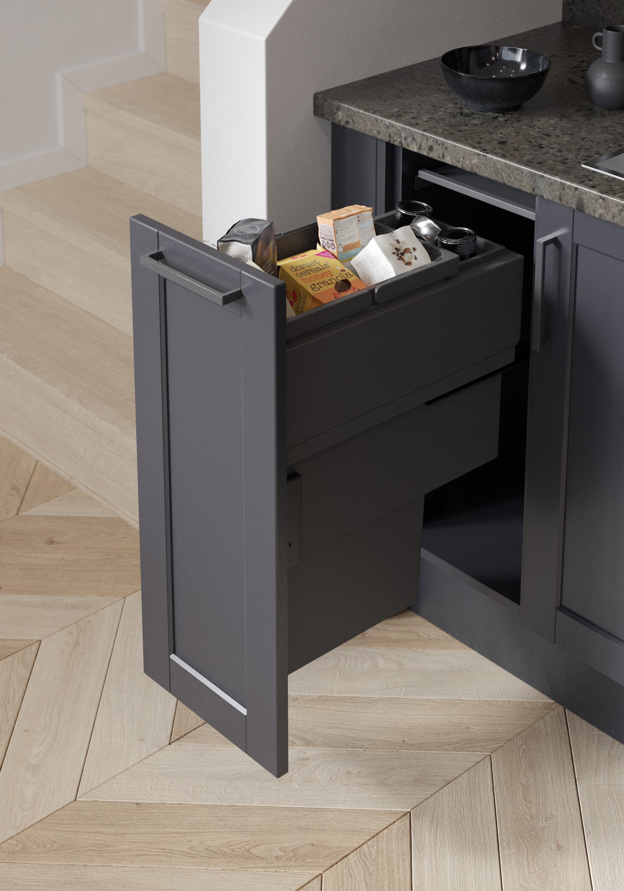 A kitchen cabinet drawer open with various packaged food items, such as cereal and snacks, inside. The cabinet is dark-colored with a sleek handle, situated next to stairs and under a countertop.
