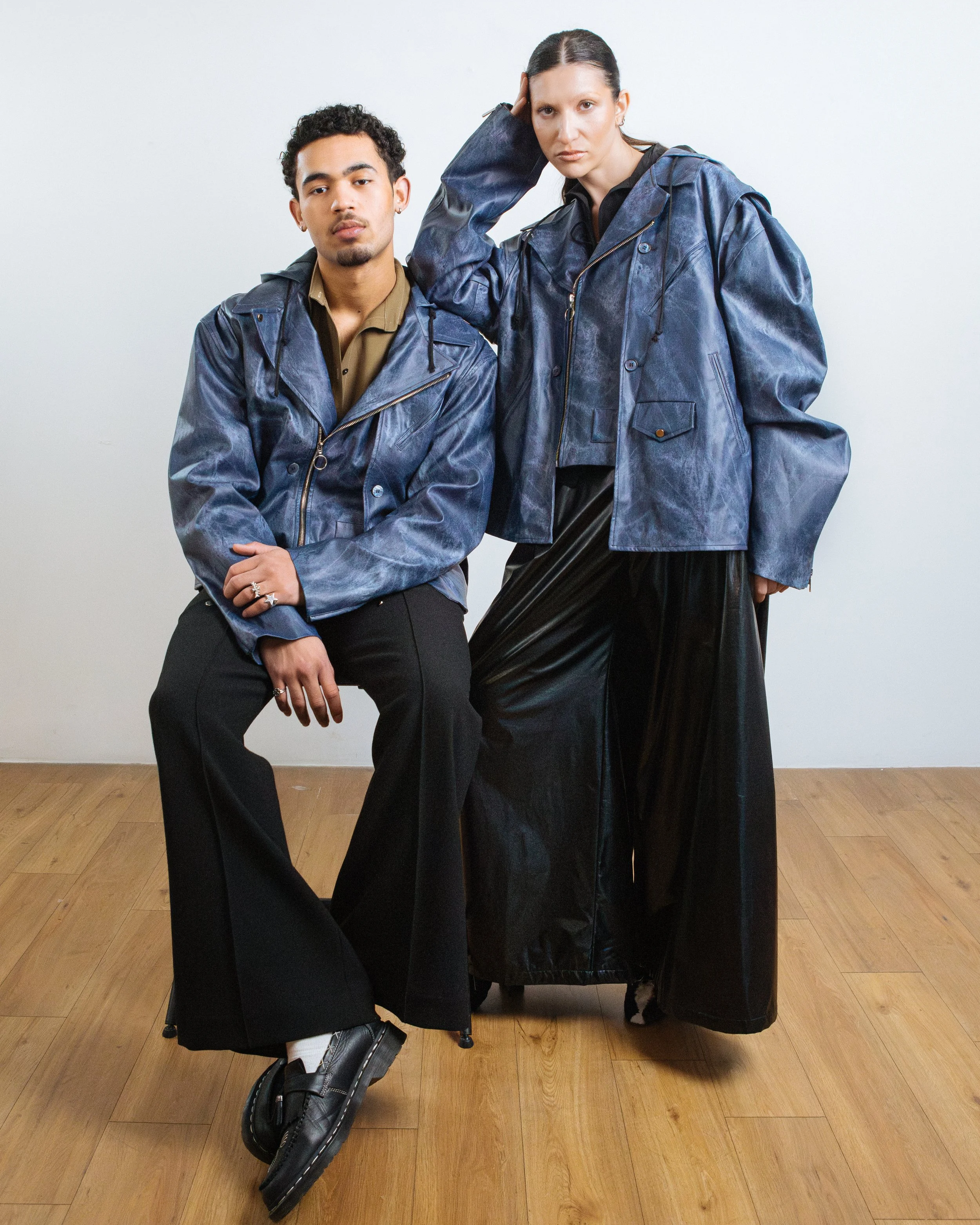 Two models wearing blue leather jackets, one sitting on a chair and the other standing with a hand on her head, against a plain white background.