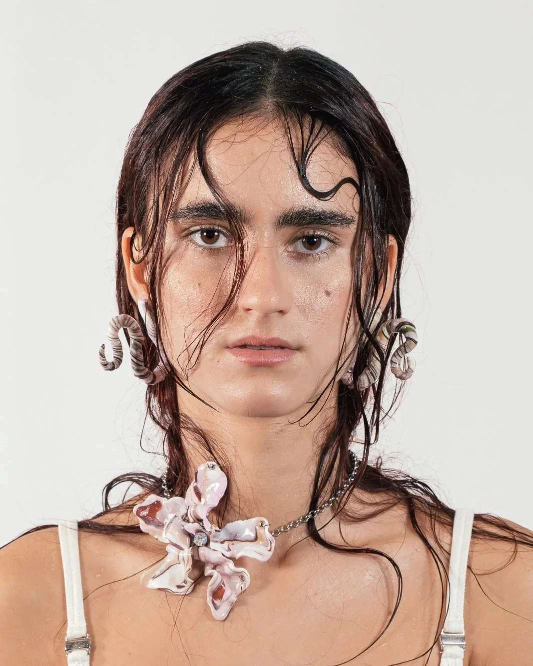 A woman with wet, dark hair, wearing large earrings and a necklace with a butterfly pendant, against a plain white background.
