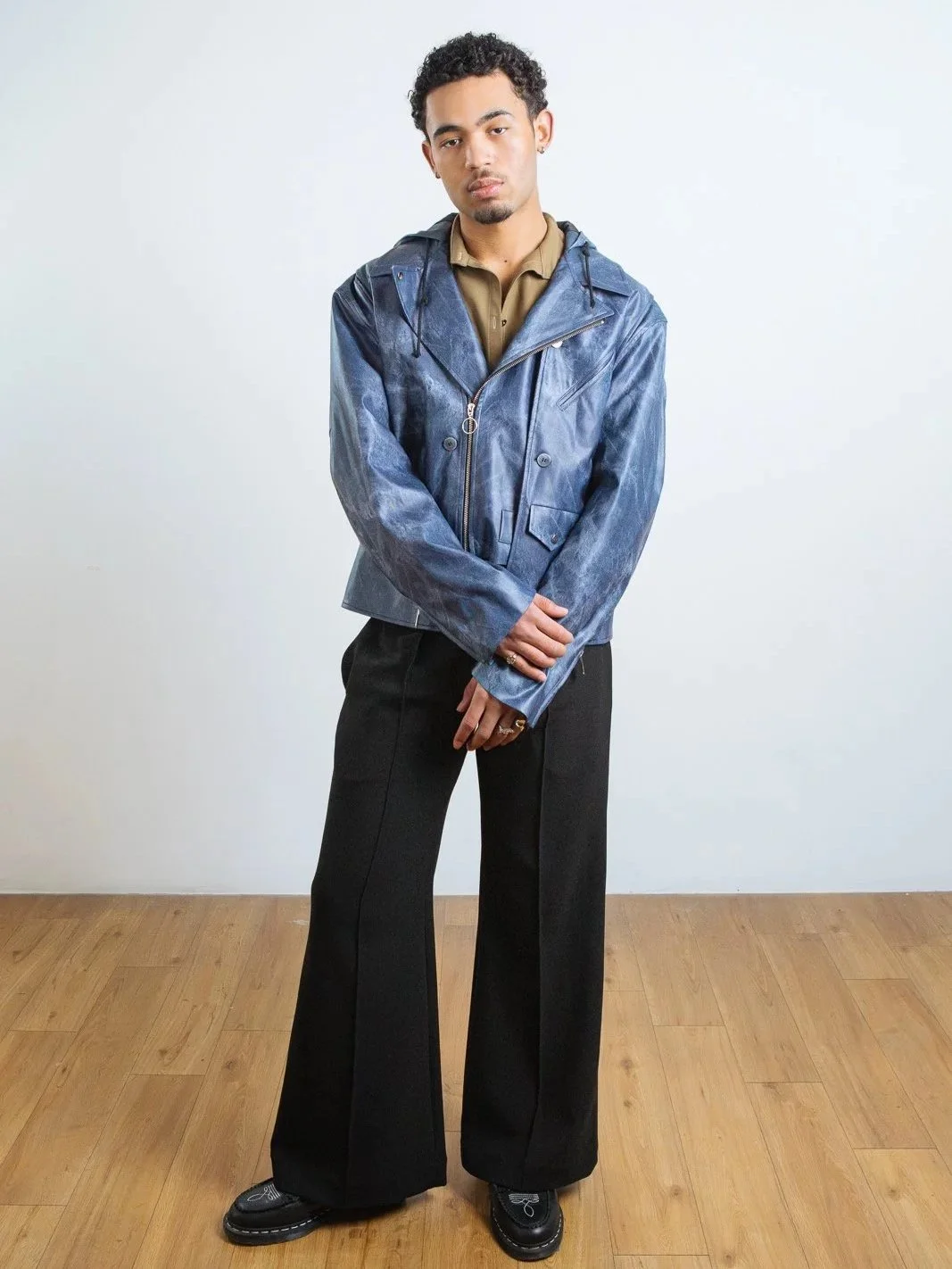 A person wearing a blue zip-up jacket, beige shirt, black wide-leg pants, and black shoes standing on a wooden floor.