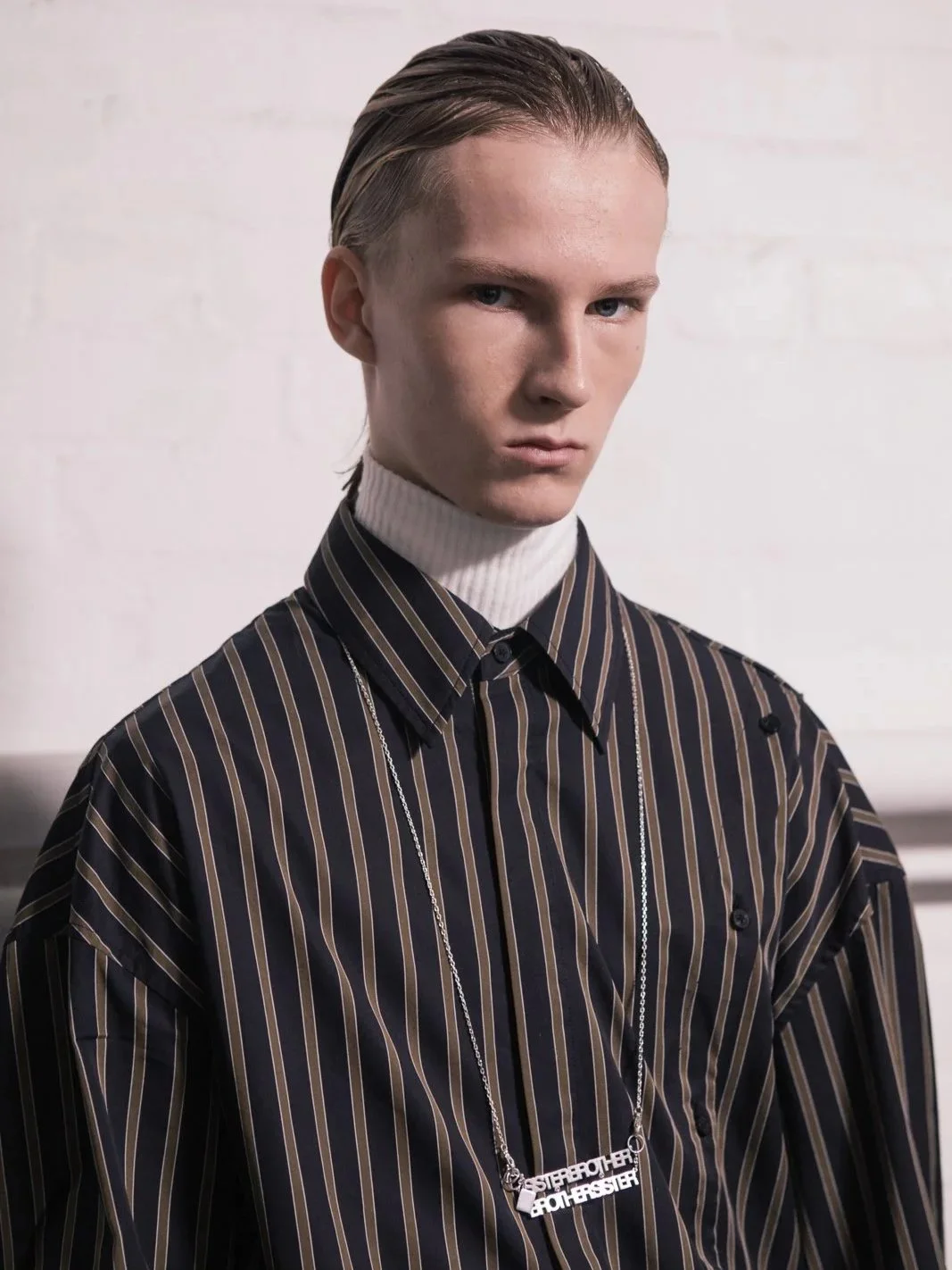 Person wearing a black pinstriped shirt and knitted turtleneck with a pendant necklace, against a white brick wall.