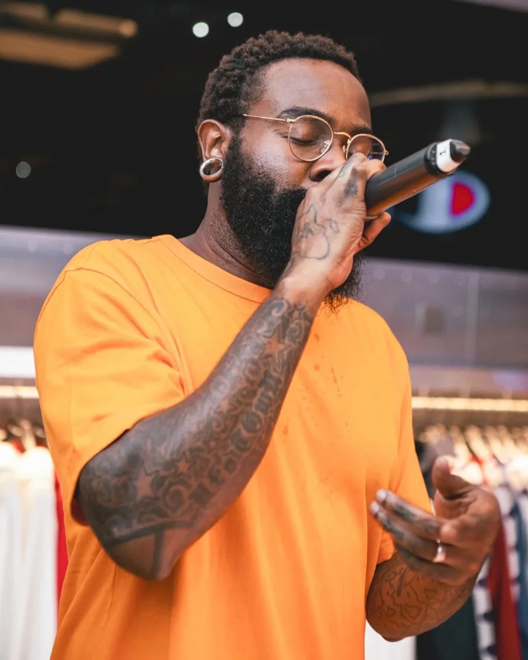 Person with tattoos and glasses singing or speaking into a microphone.