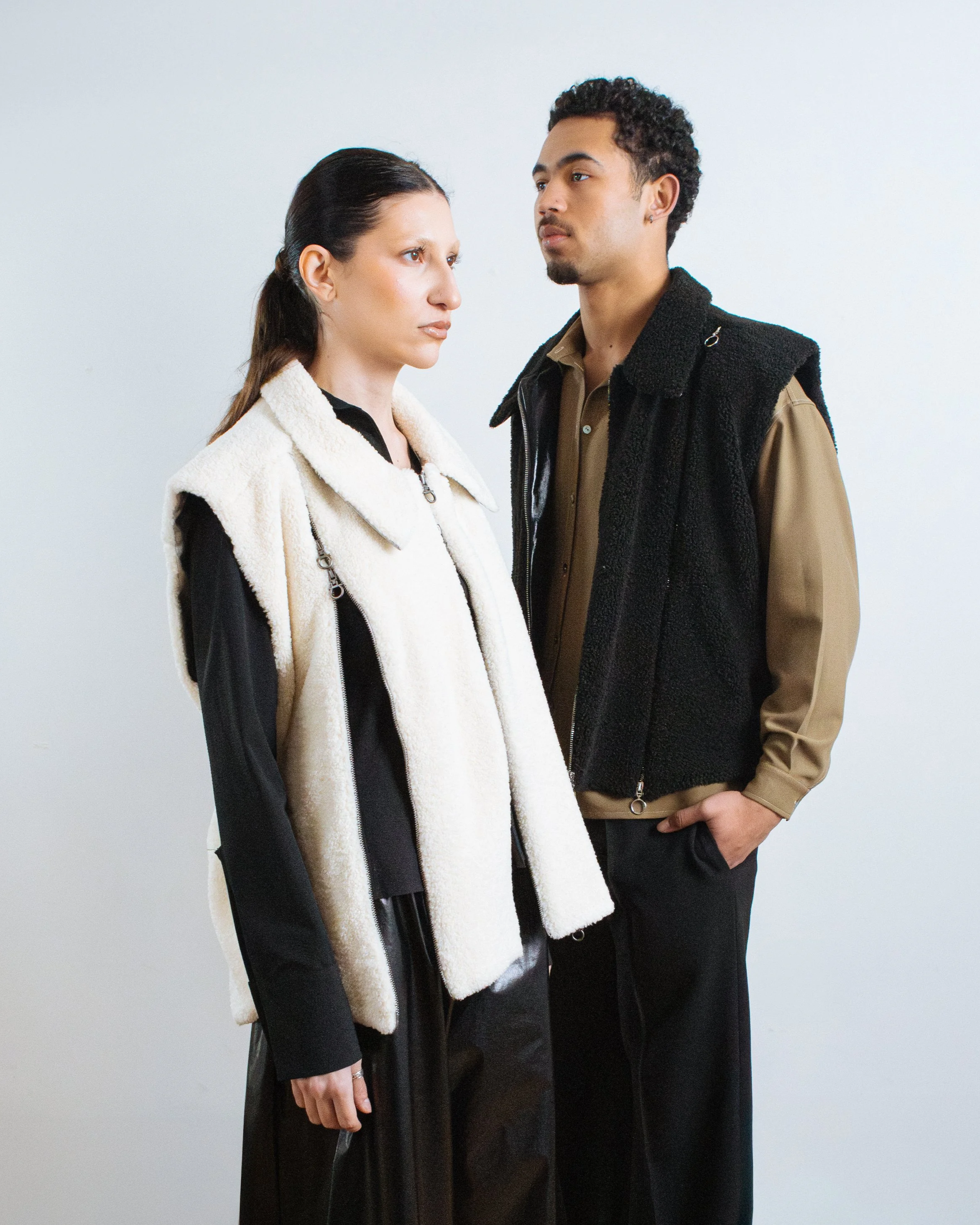A woman and a man standing face-to-face against a plain background, both looking serious. The woman has long dark hair, wearing a black outfit with a white vest. The man has curly hair, wearing a brown shirt with a black vest.