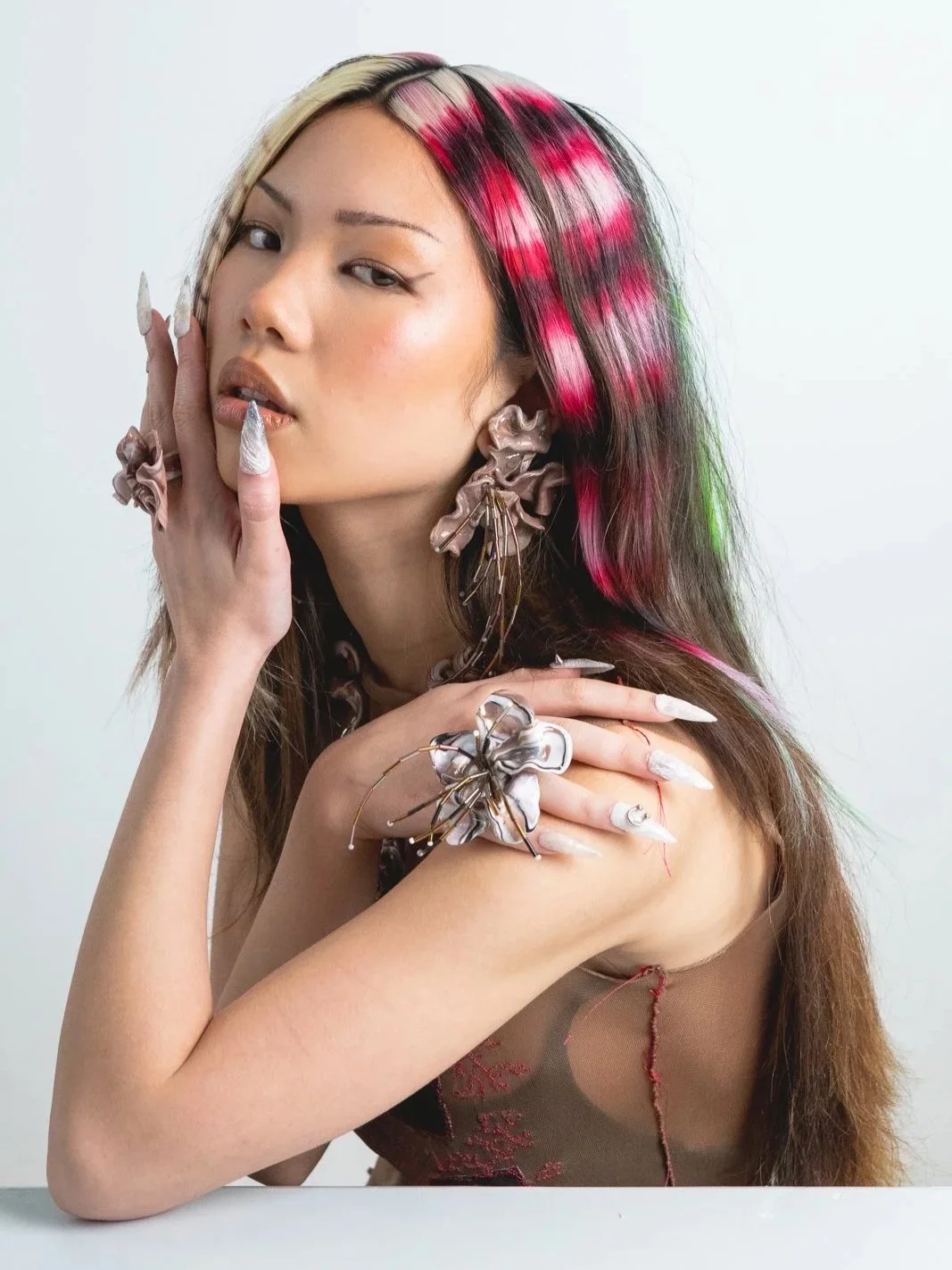 A woman with multi-colored hair featuring blonde, pink, and black streaks, wearing large decorative earrings and matching jewelry, posing with her hand on her face and shoulder against a plain background.