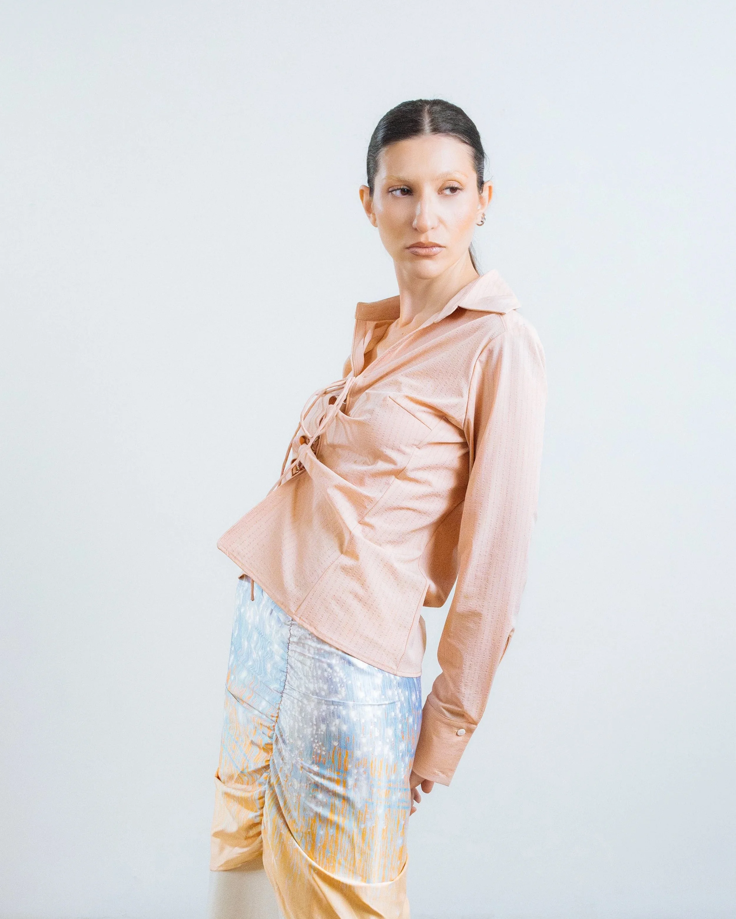 A woman with dark hair tied back, wearing a light pink blouse and colorful pants, standing against a plain white background.