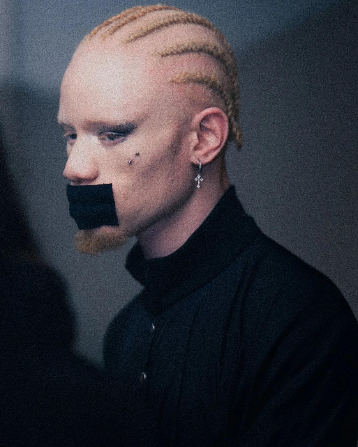 Close-up of a man with a braided blonde mohawk, multiple facial piercings, and earrings, wearing a black shirt with a black tape over his mouth.