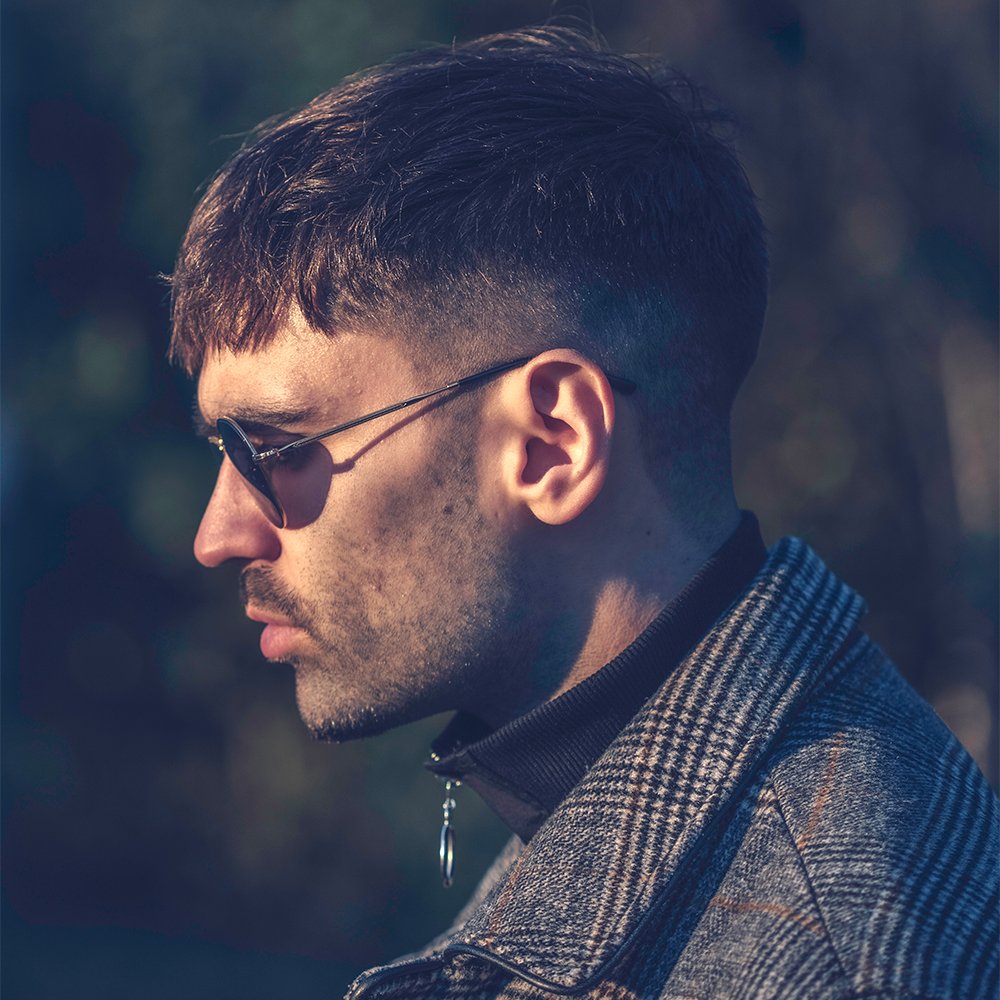 Side profile of a man wearing sunglasses and a plaid coat in natural light.