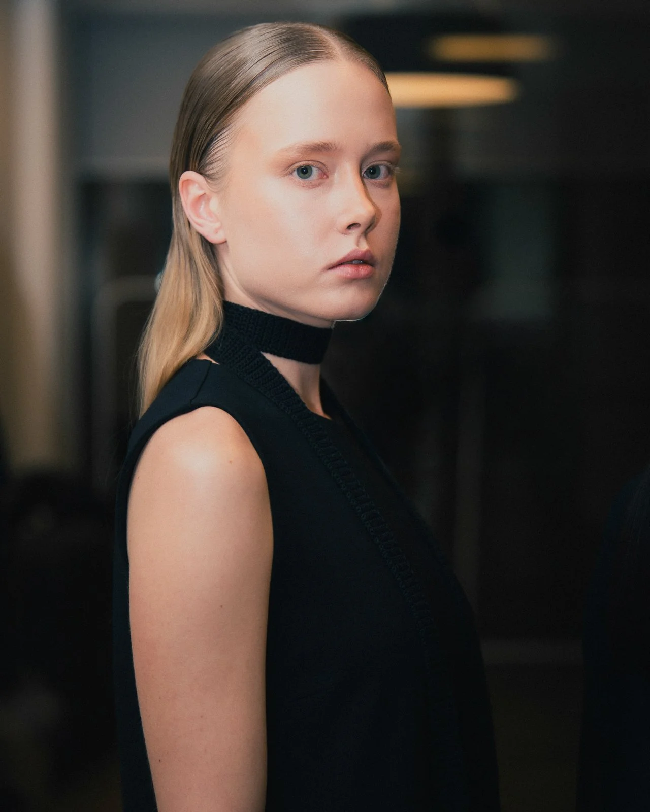A young woman with blonde hair and blue eyes looking over her shoulder, wearing a sleeveless black top and a black choker.