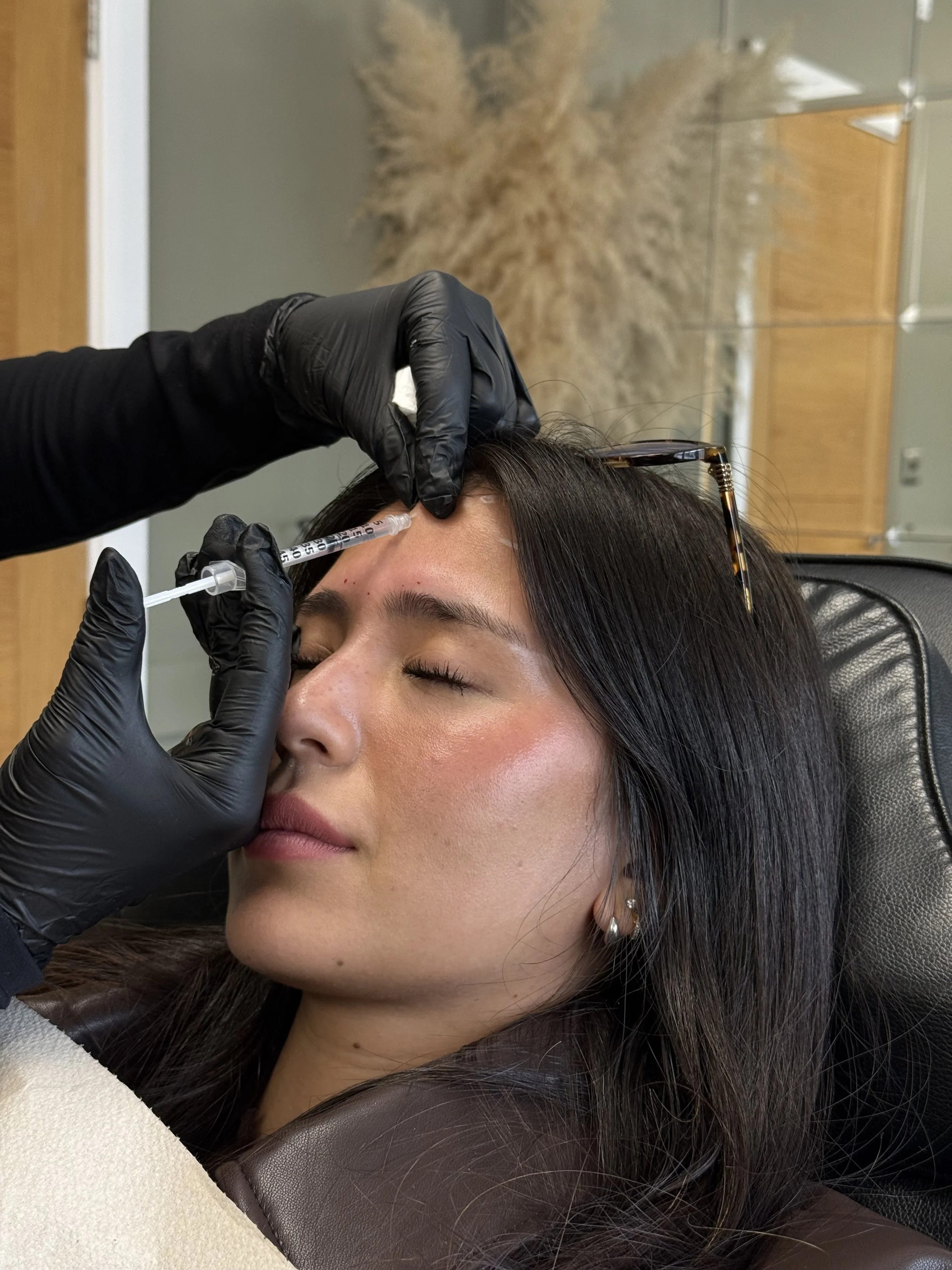A woman with closed eyes is receiving an injection in her forehead from a medical professional wearing black gloves. The woman has long dark hair, earrings, and a pair of glasses resting on her head.