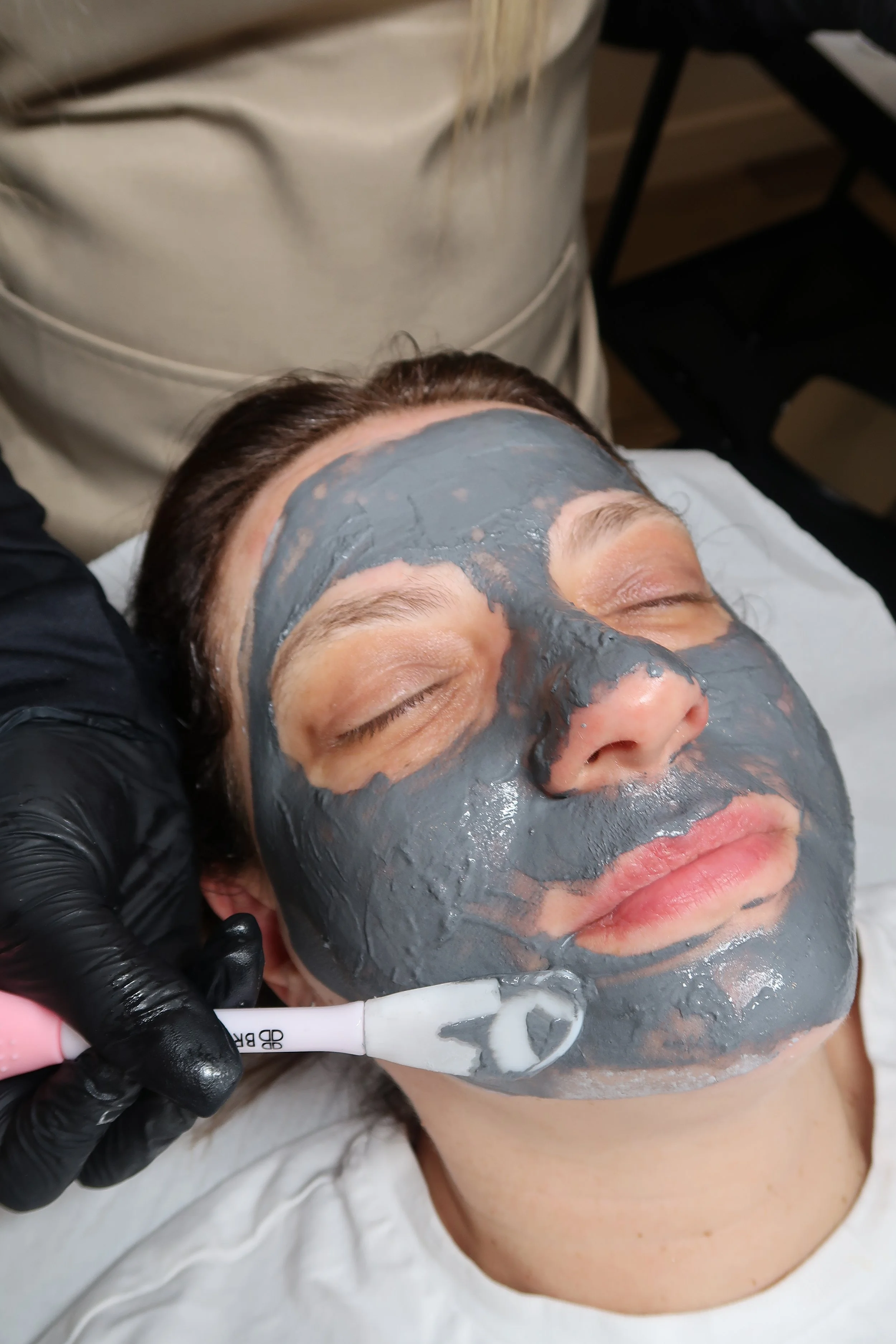 A person receiving a facial treatment with a grey facial mask applied to their face, lying on a treatment bed with eyes closed, while a technician applies the mask using a brush.