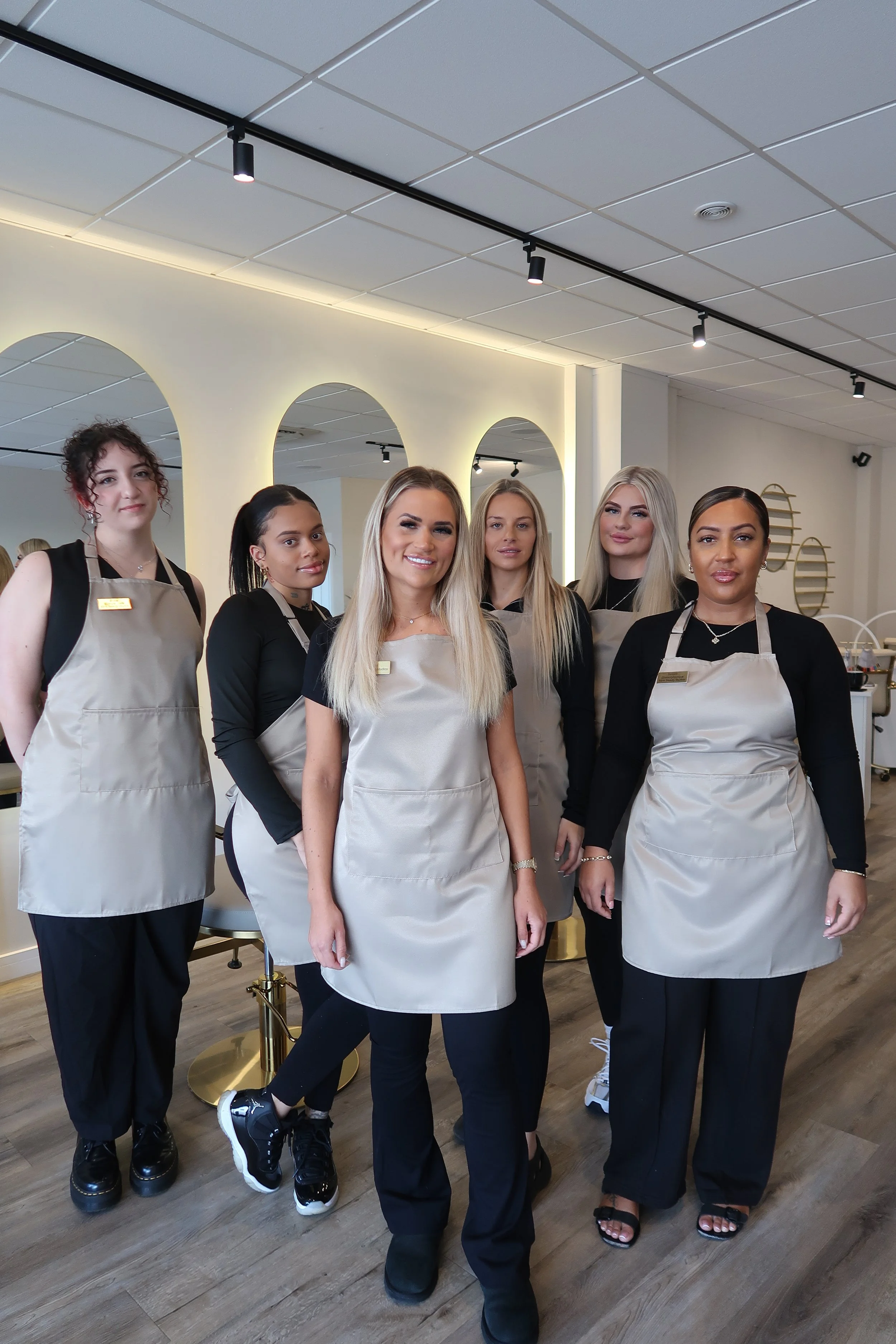 A group of six women standing indoors, all wearing beige aprons and black clothing, smiling at the camera.