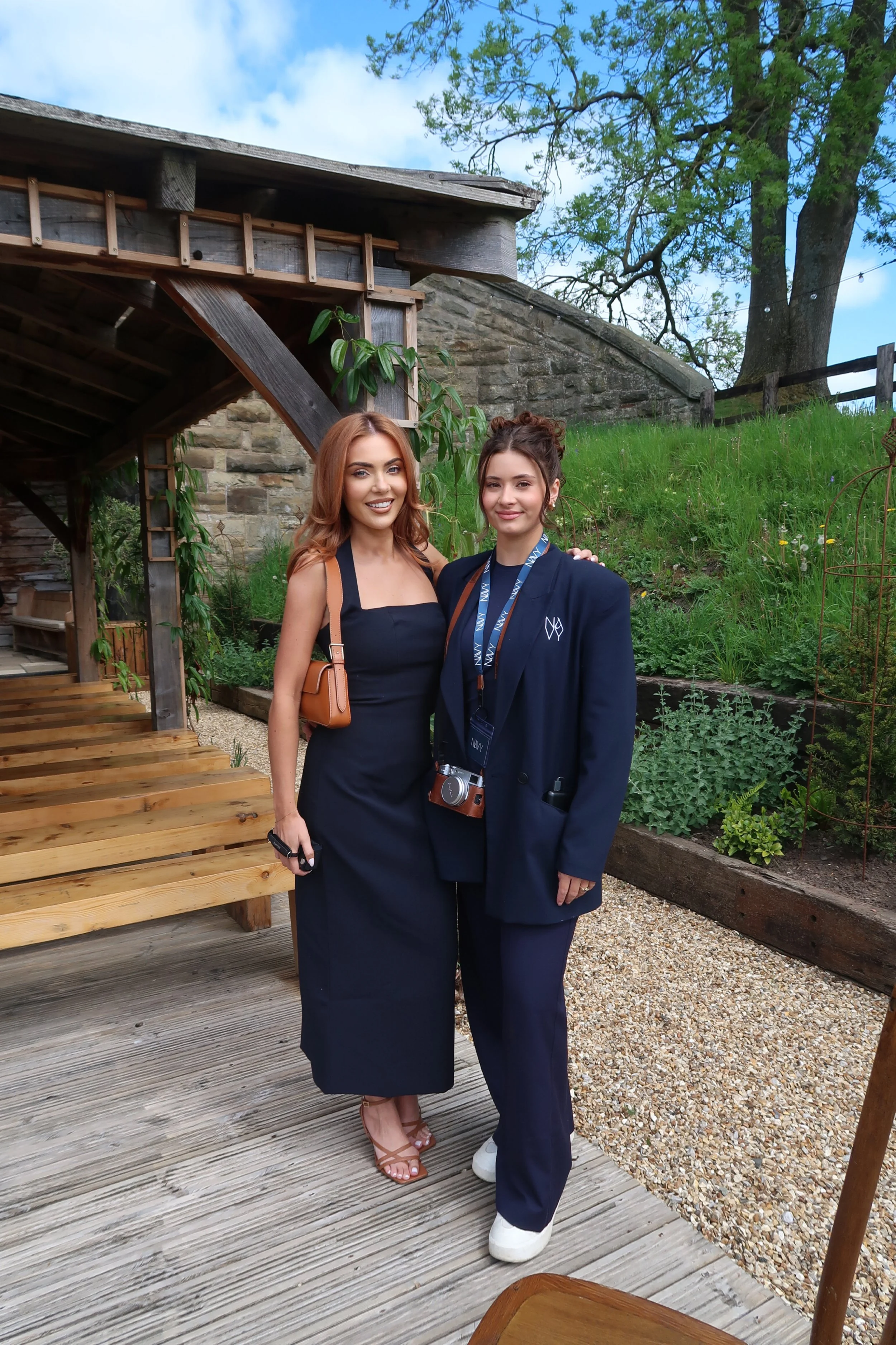 Two women standing outdoors in a garden, smiling at the camera. One woman is wearing a black dress with a tan purse, and the other is in a navy blazer and navy pants with a camera around her neck.