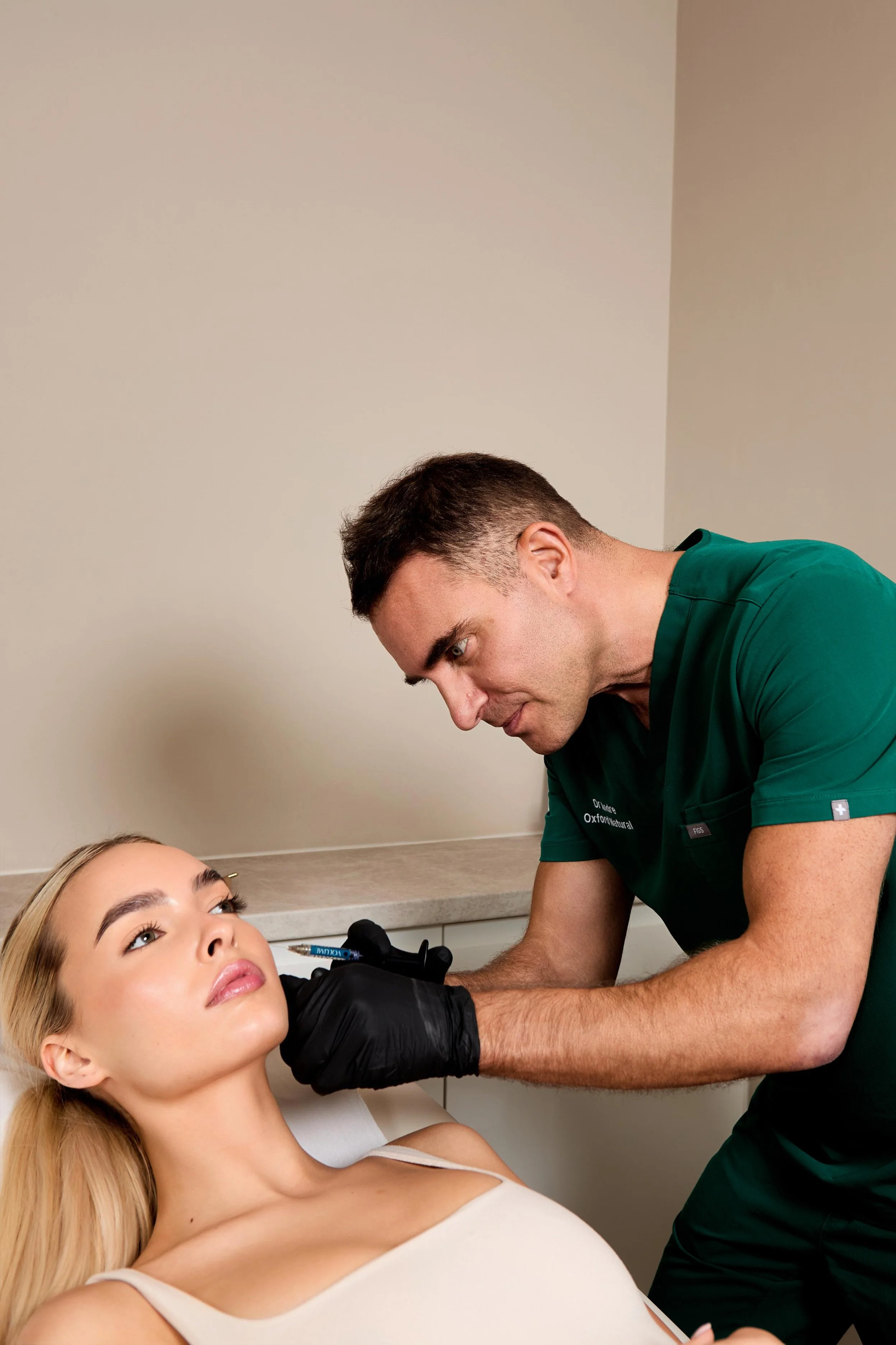 Medical professional in green scrubs and black gloves administering a cosmetic injection to a woman lying on a medical bed.