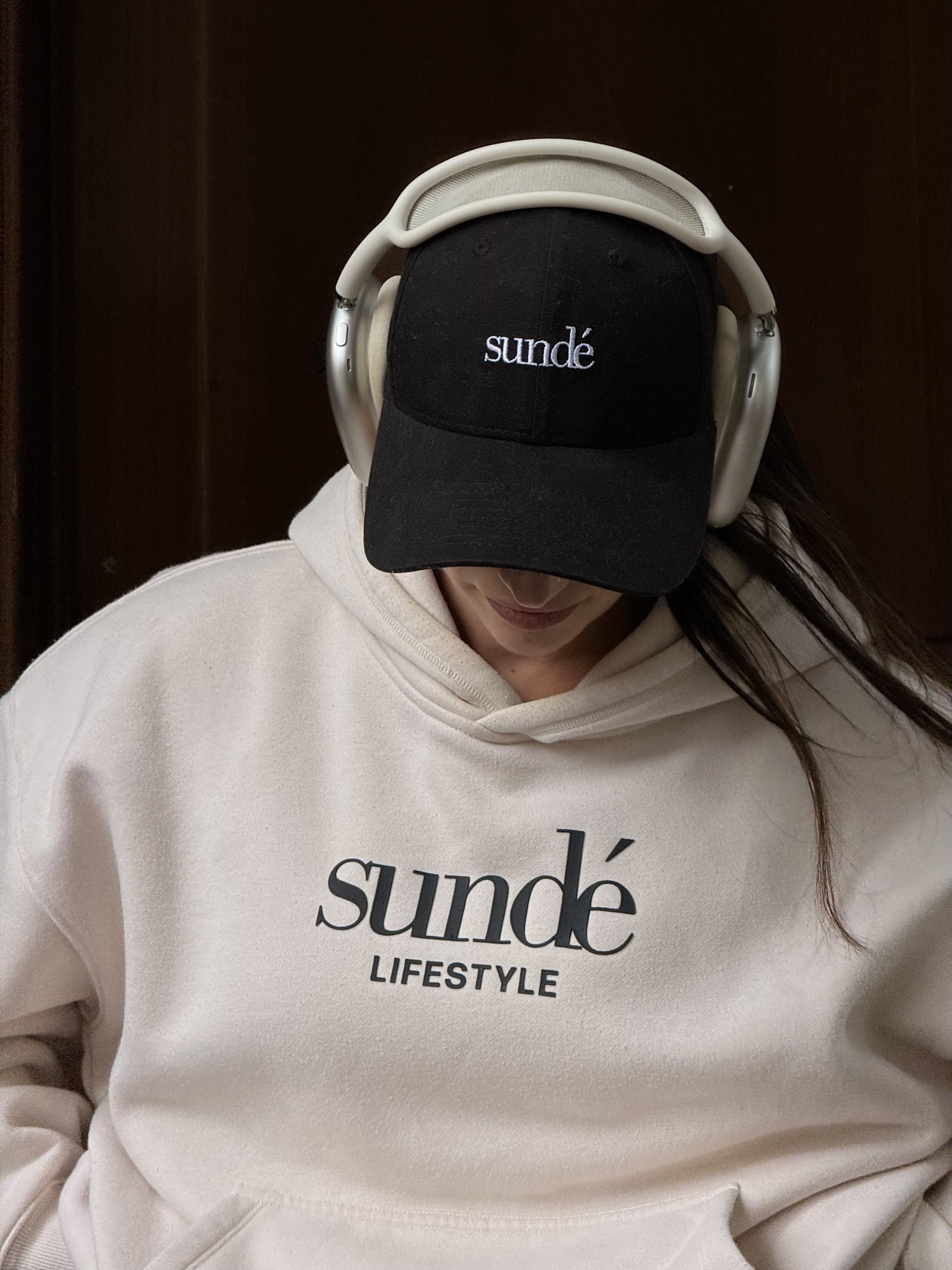 Person wearing a black baseball cap with 'sundé' embroidered on it, a white hoodie with 'sundé LIFESTYLE' printed on the front, and over-ear headphones, looking downward, against a dark background.