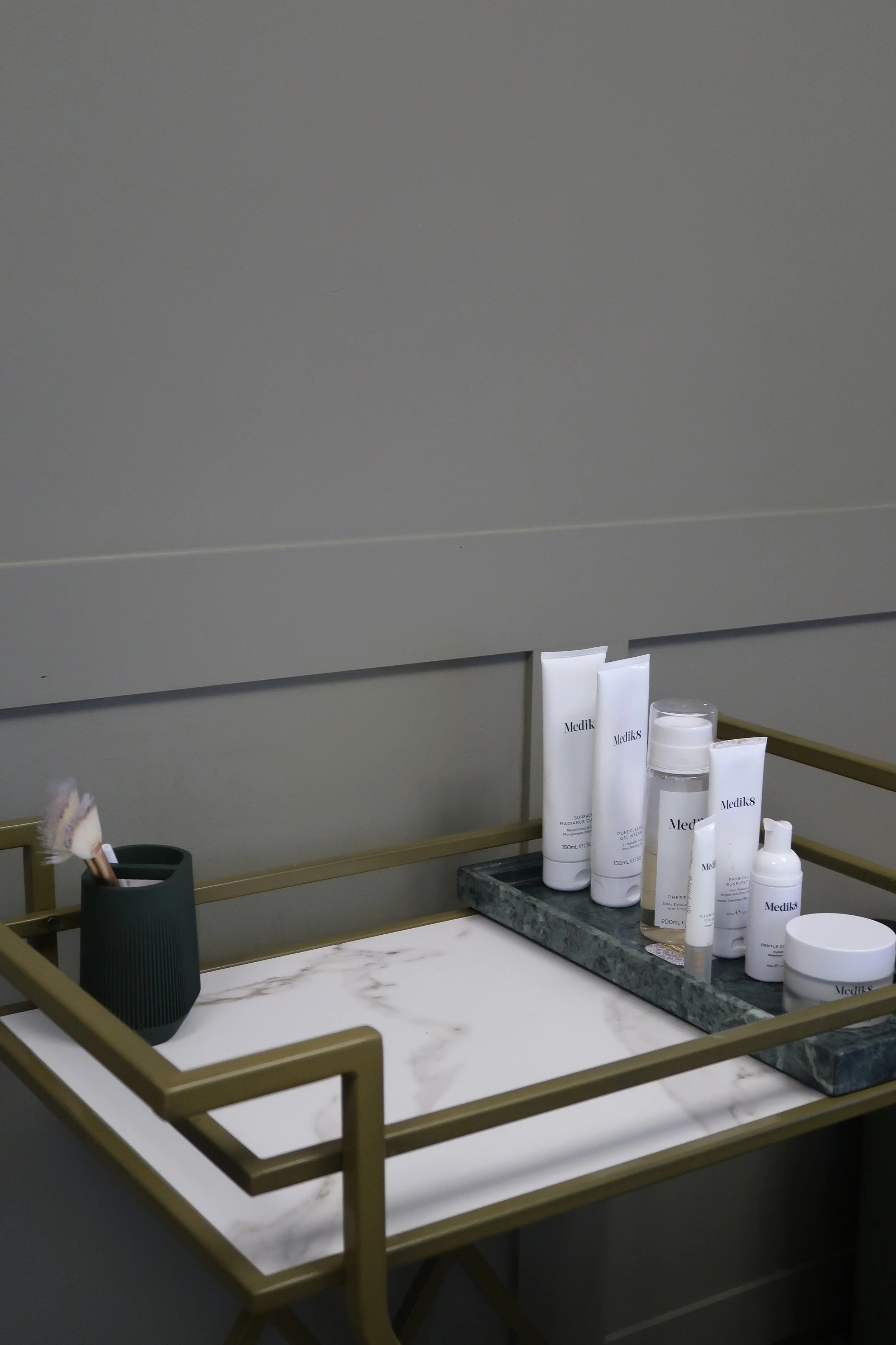 A display tray with skincare products on a marble surface, demonstrating a minimalist setup with a gray wall background. The products are labeled 'Medik8' and include creams, serums, and sprays.