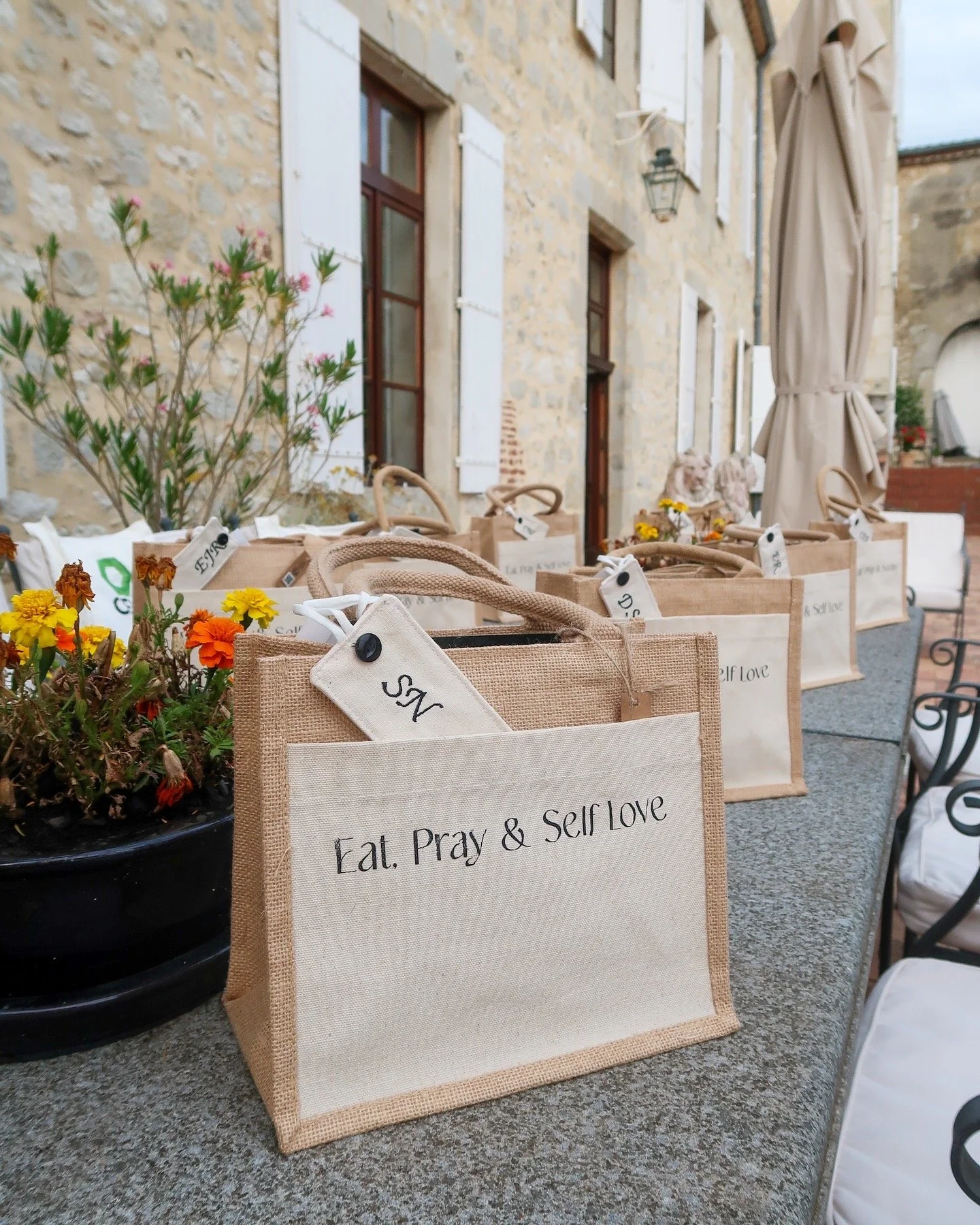 Multiple beige fabric tote bags with the phrase 'Eat, Pray & Self Love' on them, arranged on a table next to potted flowers outside a stone building.