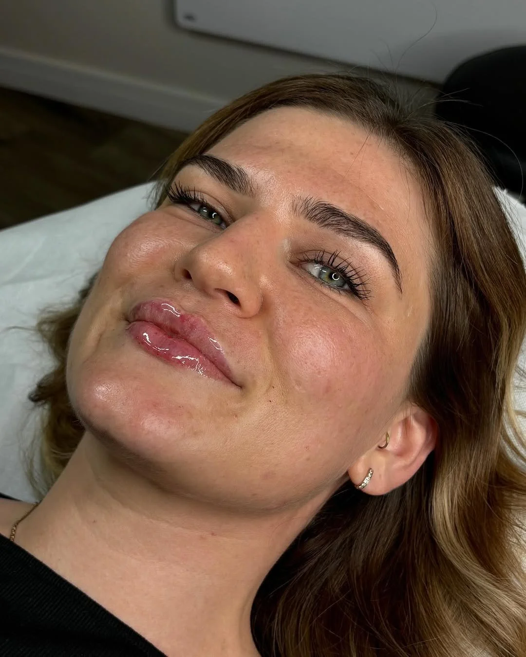A woman lying on a white pillow with shiny, full lips and well-groomed eyebrows, smiling slightly and looking at the camera.