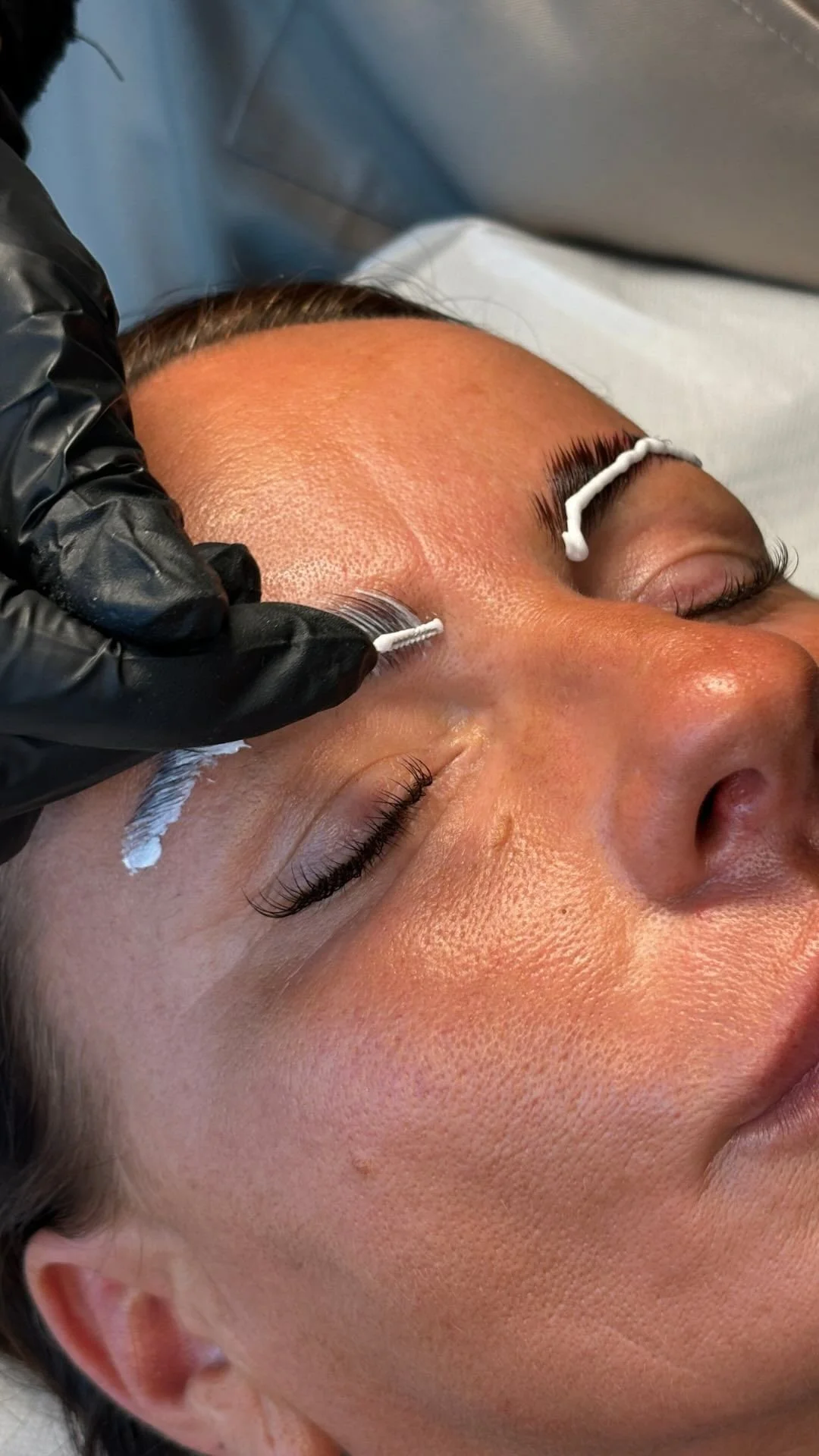 A woman receiving eyelash extension application, lying down with eyes closed, with cotton swabs placed between her eyelashes and eyelids.