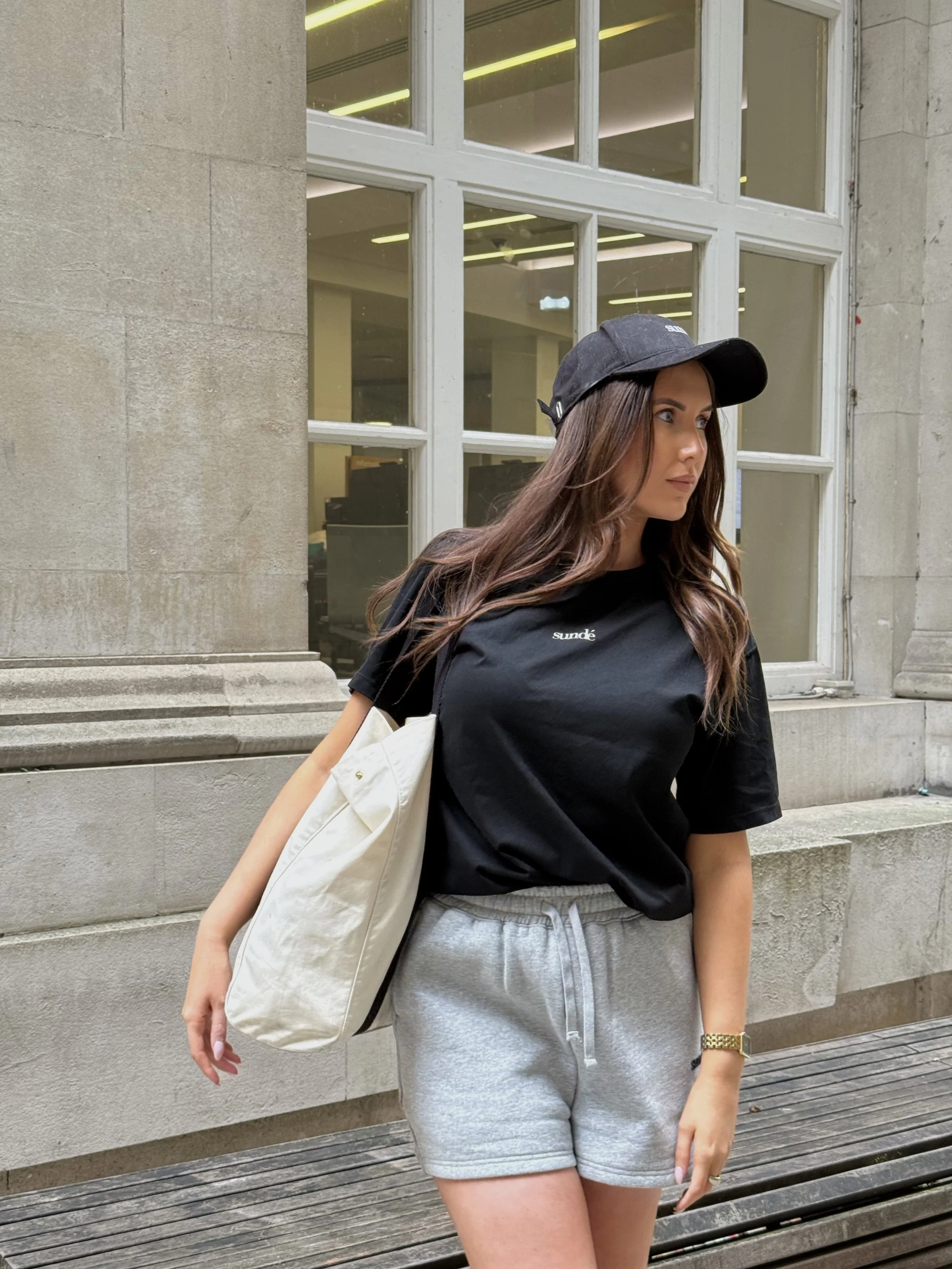 A young woman with long brown hair, wearing a black baseball cap, black t-shirt, and gray shorts, standing outdoors near a stone building with large windows, carrying a beige tote bag over her shoulder.
