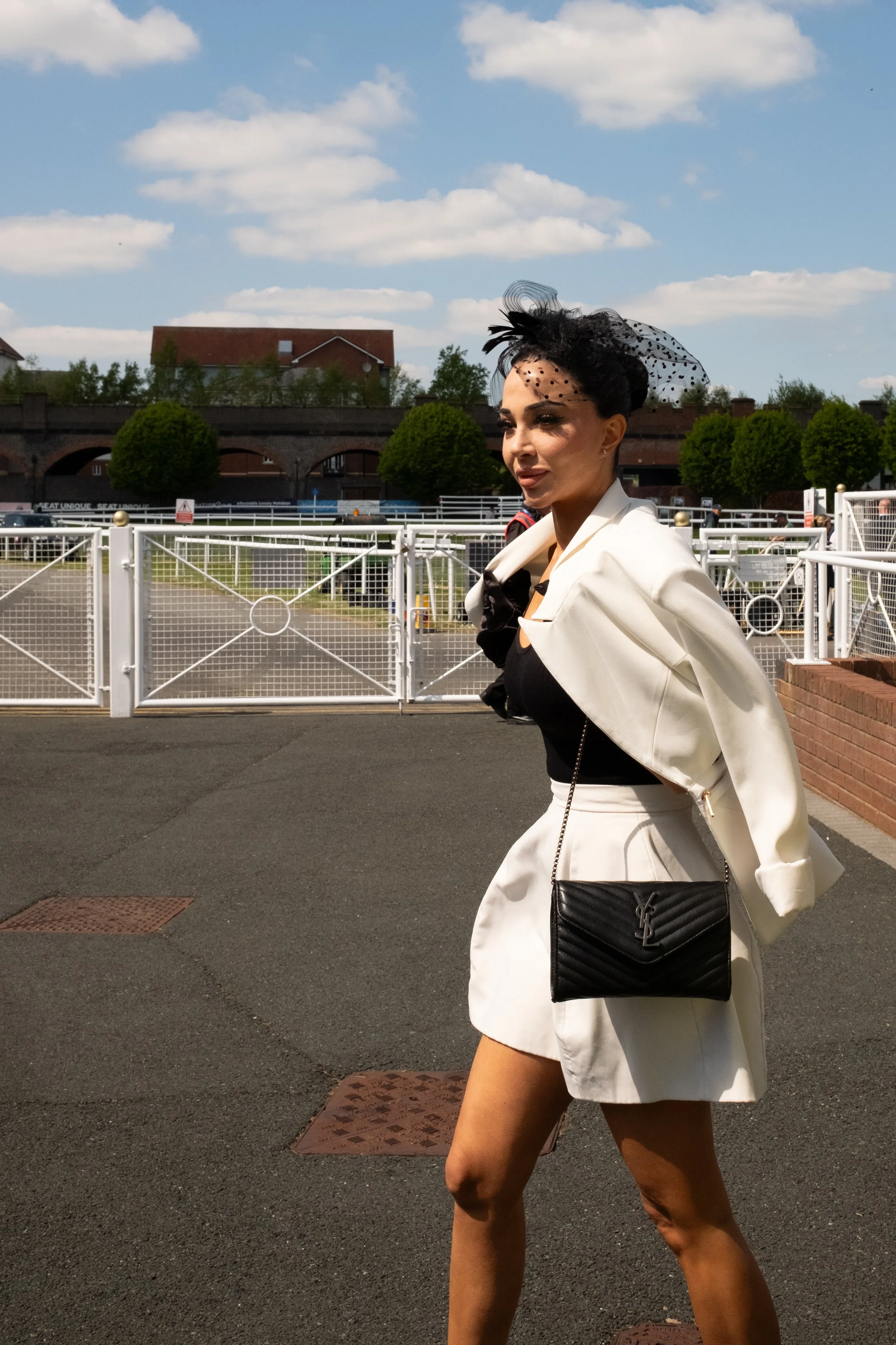 A woman wearing a white blazer, matching white skirt, and a black top, carrying a black Yves Saint Laurent purse with a chain strap, walking outdoors near a white fence and brick wall under a partly cloudy sky.