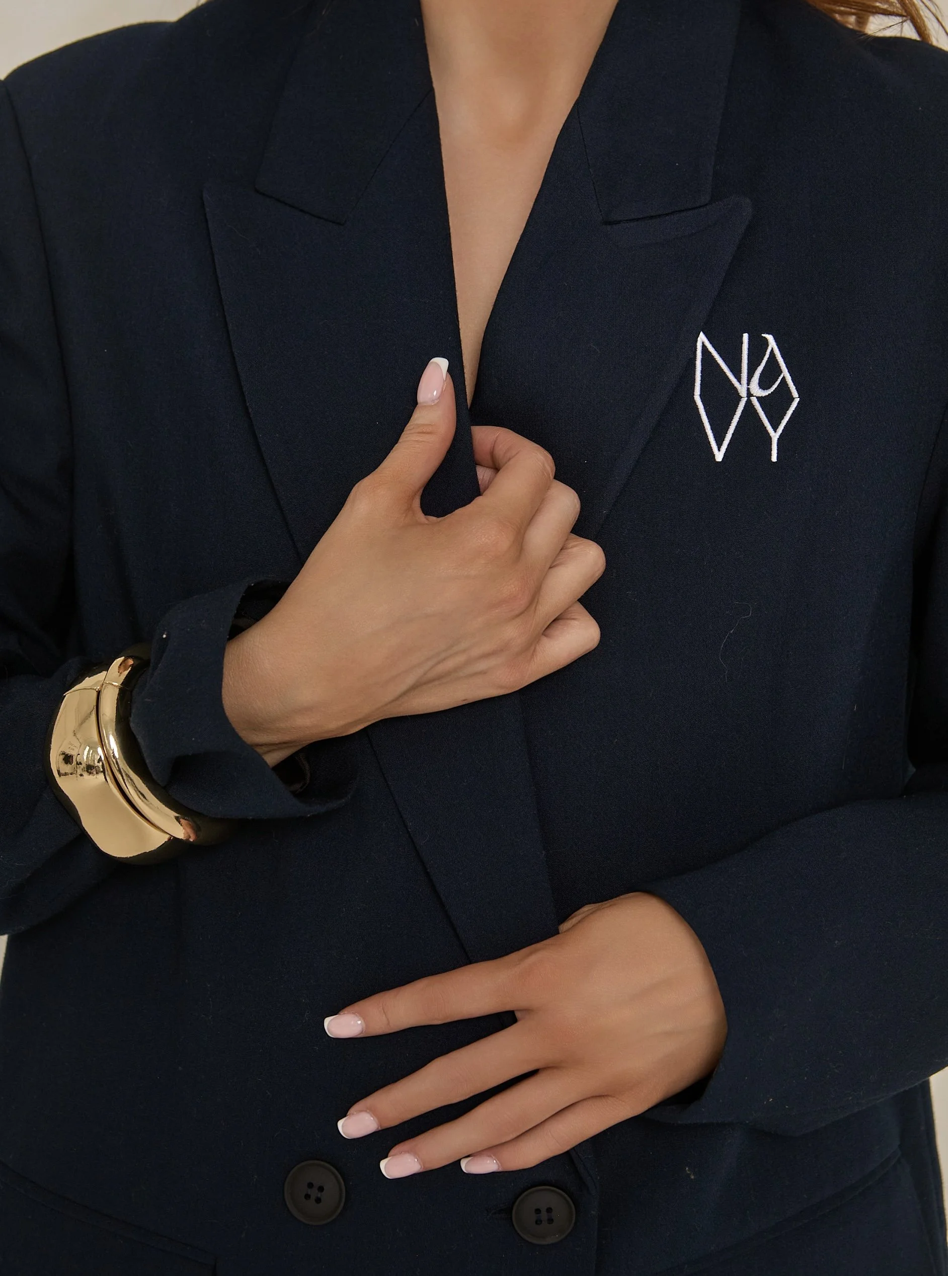 A person wearing a dark blazer with a white embroidered logo, touching their chest with one hand and pointing to their ring finger with the other. The person has a gold bracelet on their wrist and manicured nails with white tips.