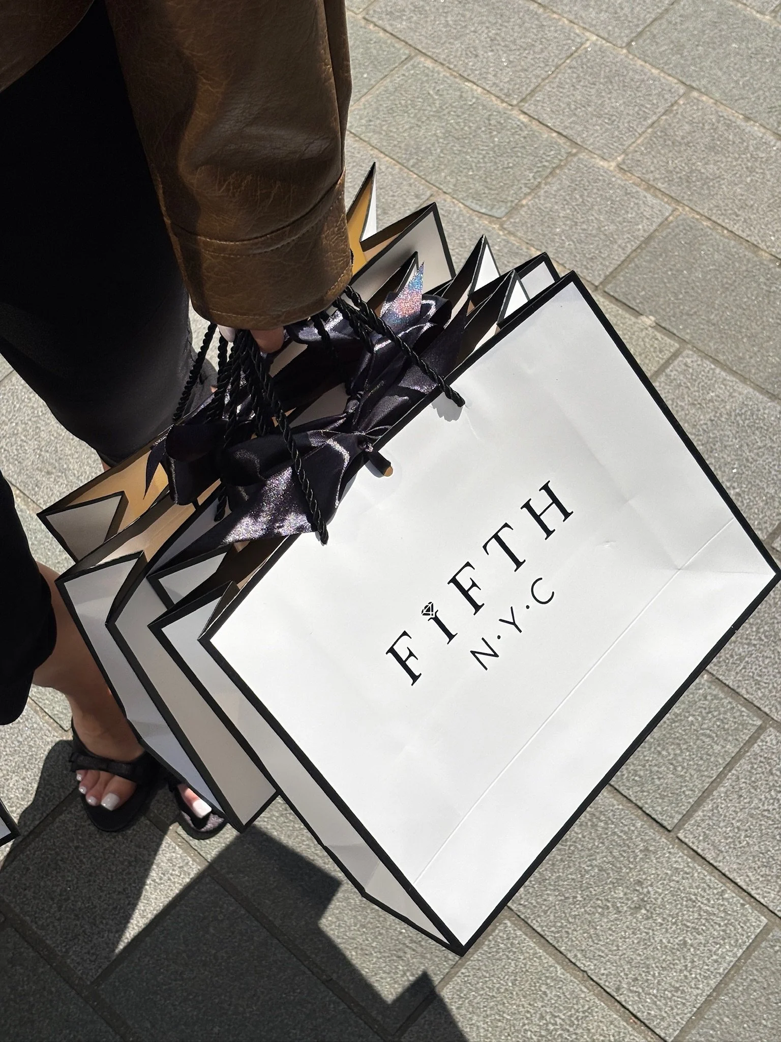 A person is holding multiple FIFTH NYC shopping bags on a sidewalk.