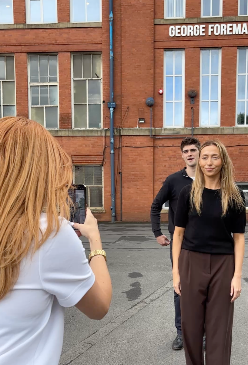 A woman with red hair is taking a photo of a man and a woman standing outside in front of a brick building labeled 'George Foreman'. The woman being photographed has long blonde hair, is wearing a black top and brown pants, and is smiling slightly. The man behind her has short dark hair and is wearing a black zip-up jacket.