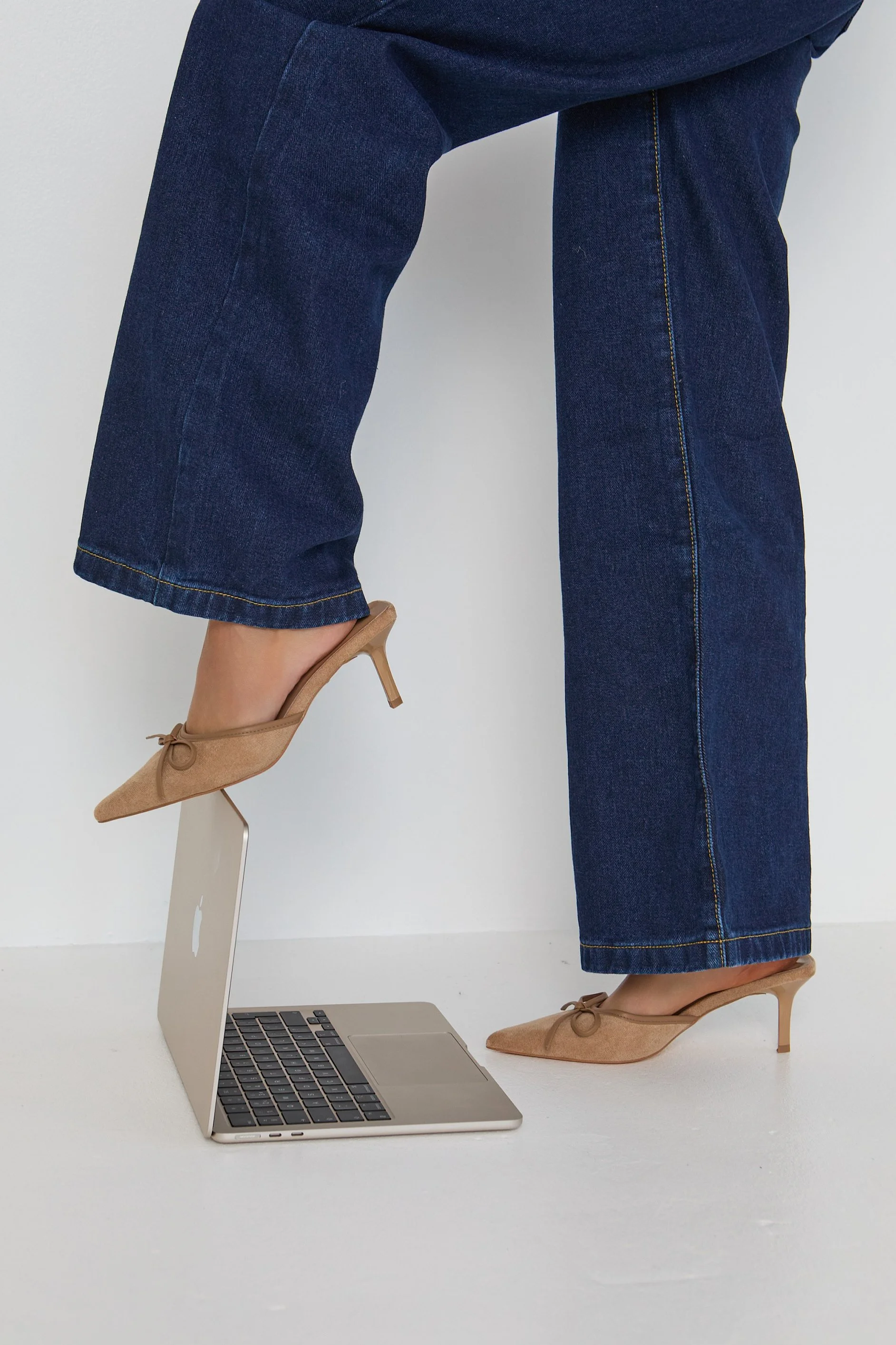 Person standing with legs apart, wearing dark blue jeans and beige high-heeled shoes, with one foot on a silver laptop keyboard, in front of a white wall.