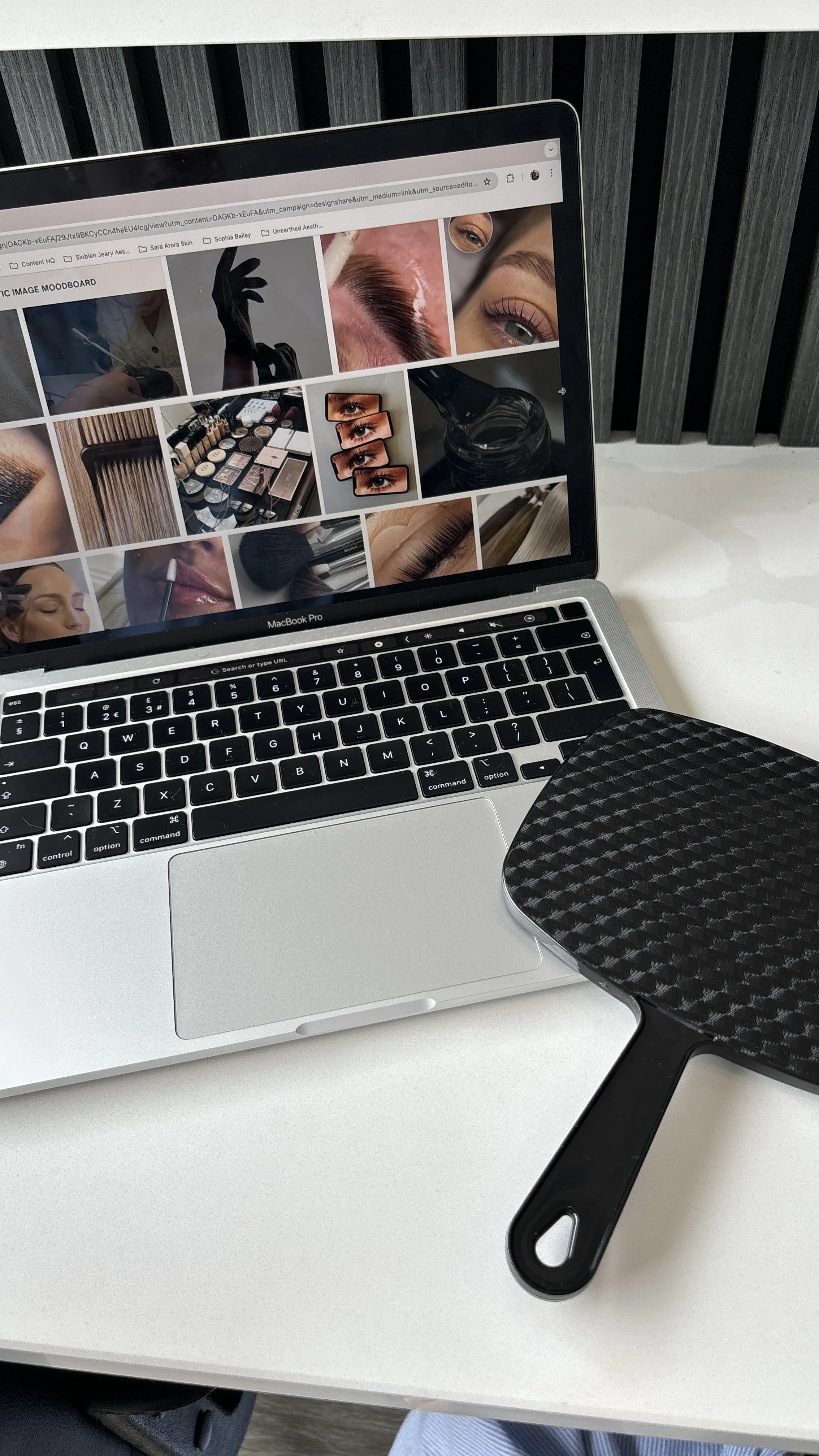 A MacBook laptop on a white desk displaying a Pinterest page with makeup, eye makeup, skincare, and beauty images. A black textured paddle brush rests on the desk beside the laptop.