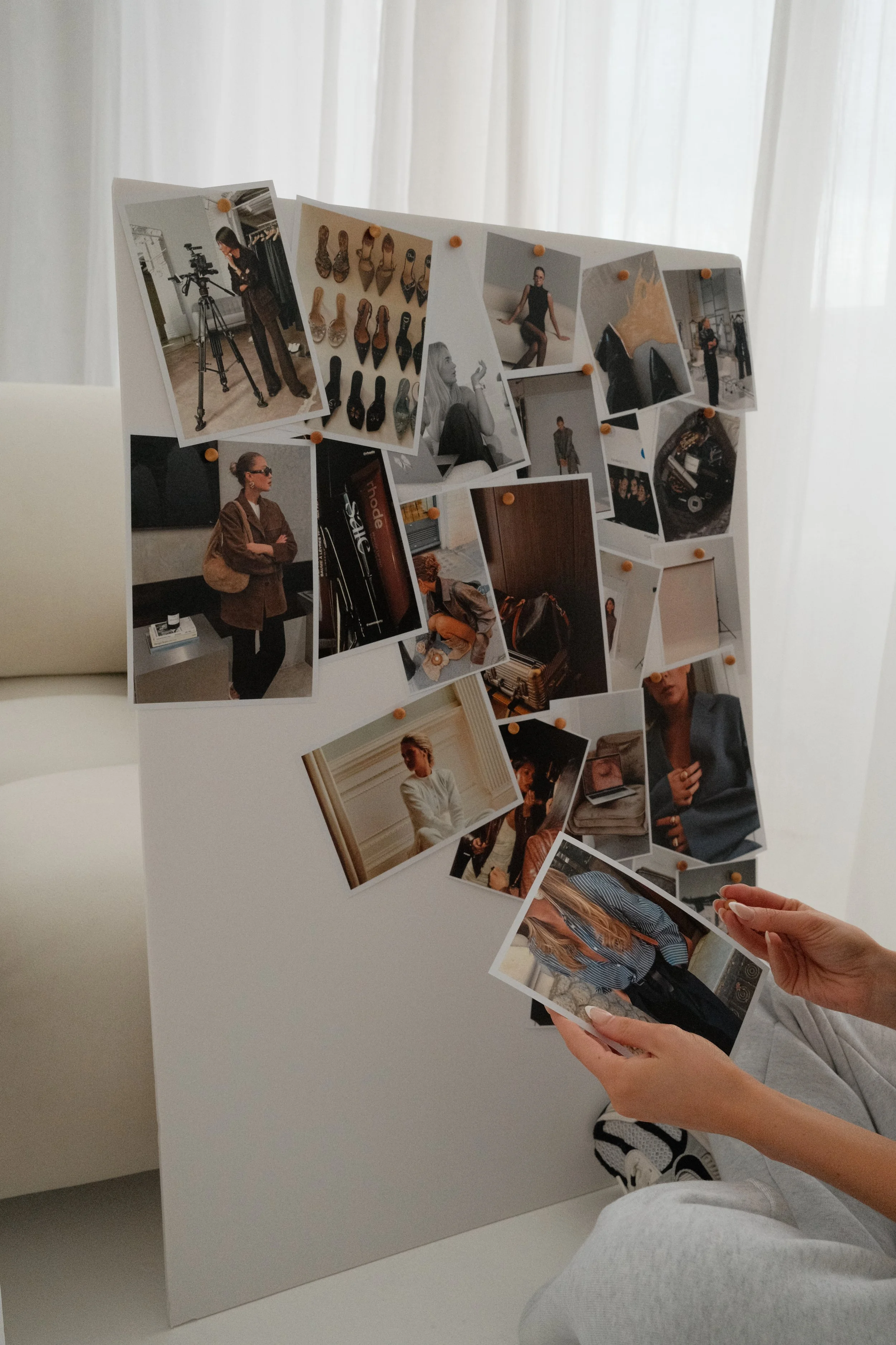 A person is looking at a display board with various photos pinned to it. The photos include images of people, shoes, and a camera setup. The person is holding a photo with a woman in a striped shirt and dark pants.